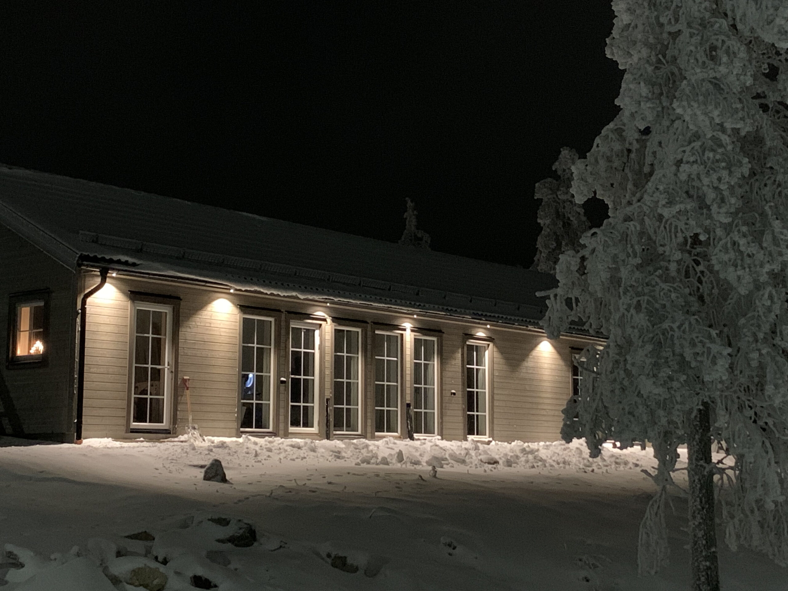 Långt ljust hus upplyst av fasadbelysning en vinterkväll, med snö på marken och snötäckta träd i förgrunden.