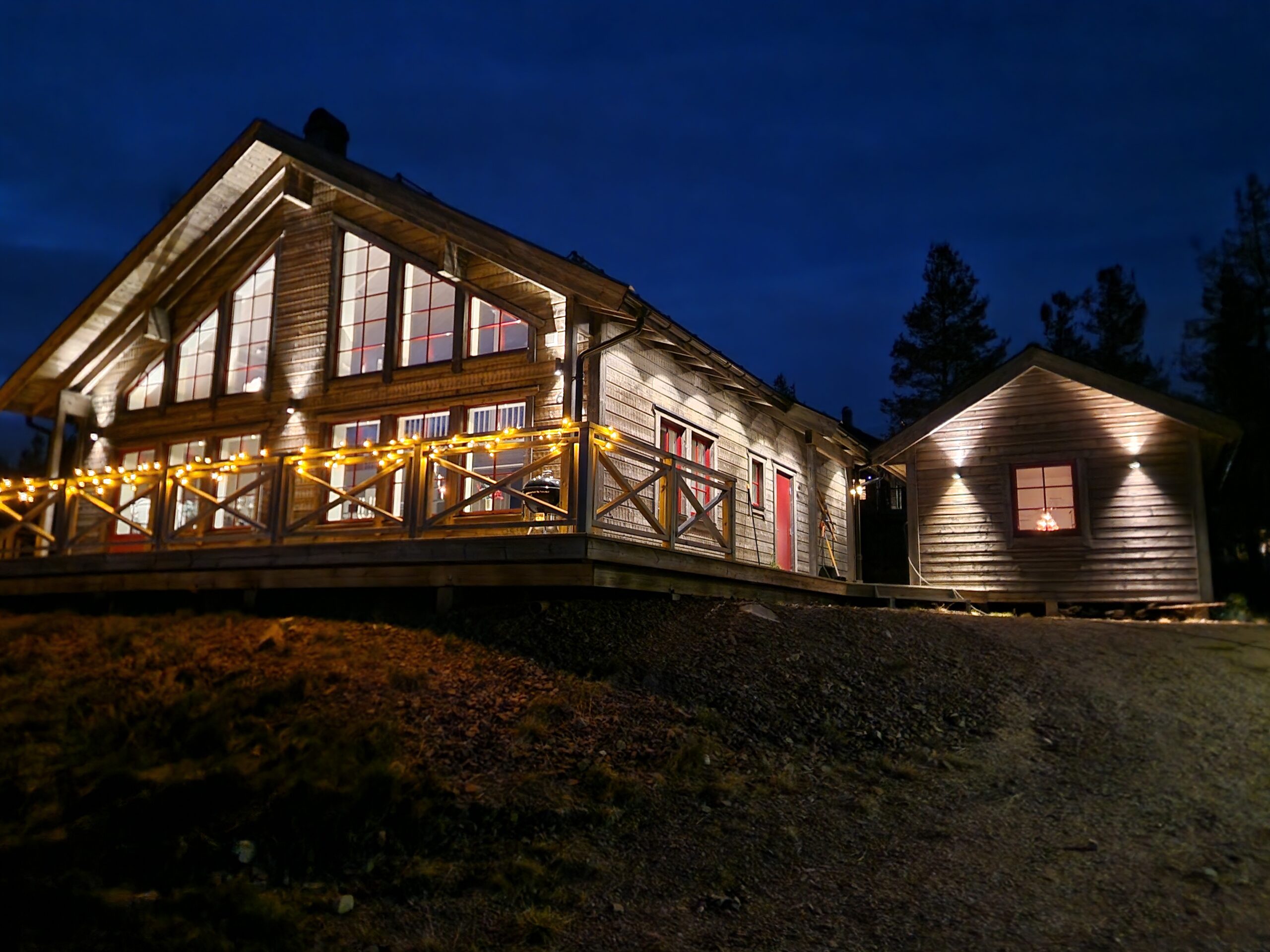 En upplyst trästuga med stora fönster och altan, fotograferad i skymningen med mörkblå himmel i bakgrunden.