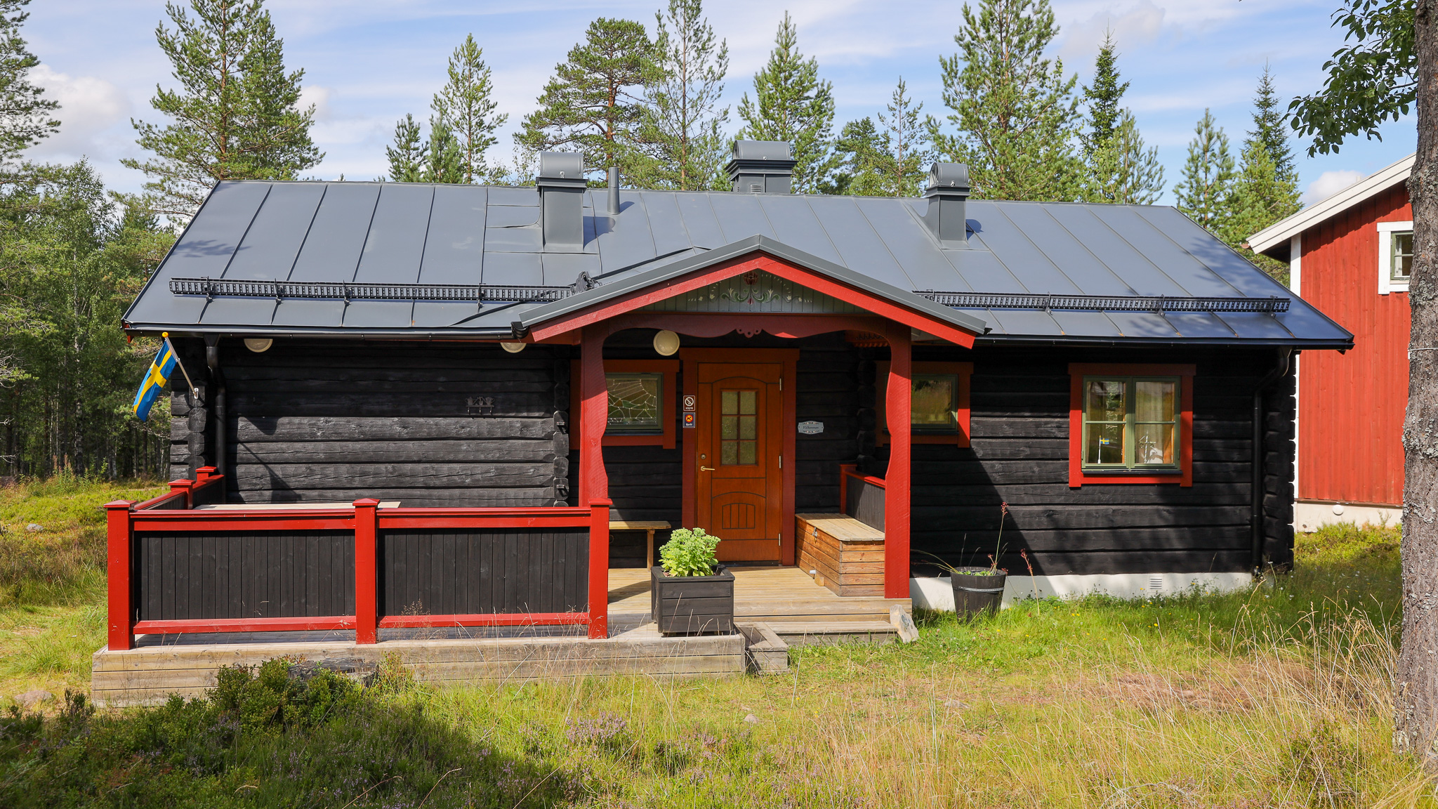 Mörk trästuga med rött takutsprång och röda räcken på altanen. Entré med trätrappa och blomkruka framför dörren. Omgiven av gräs och tallskog, med ett rött hus i bakgrunden.