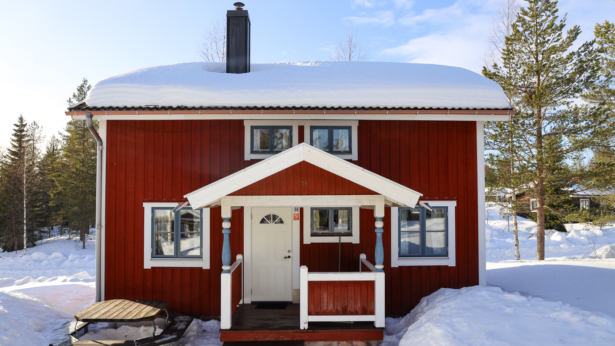 Rött trähus med vit snickarglädje och snötäckt tak, omgivet av snö och tallar under klarblå himmel.