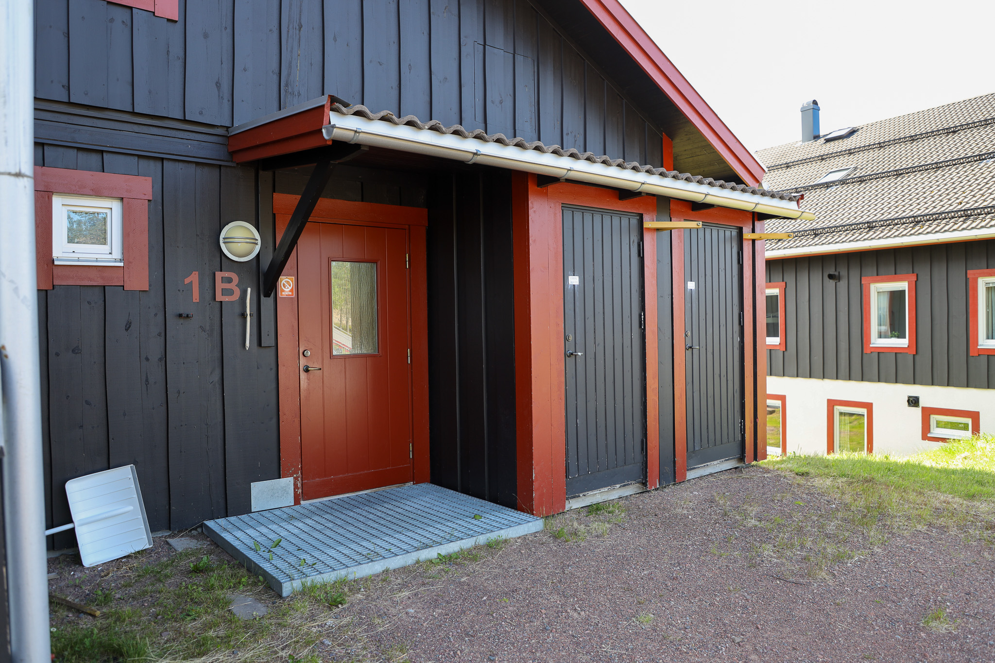 En mörk träbyggnad med röd dörr och detaljer, liten takskyddad entré och grus framför huset.