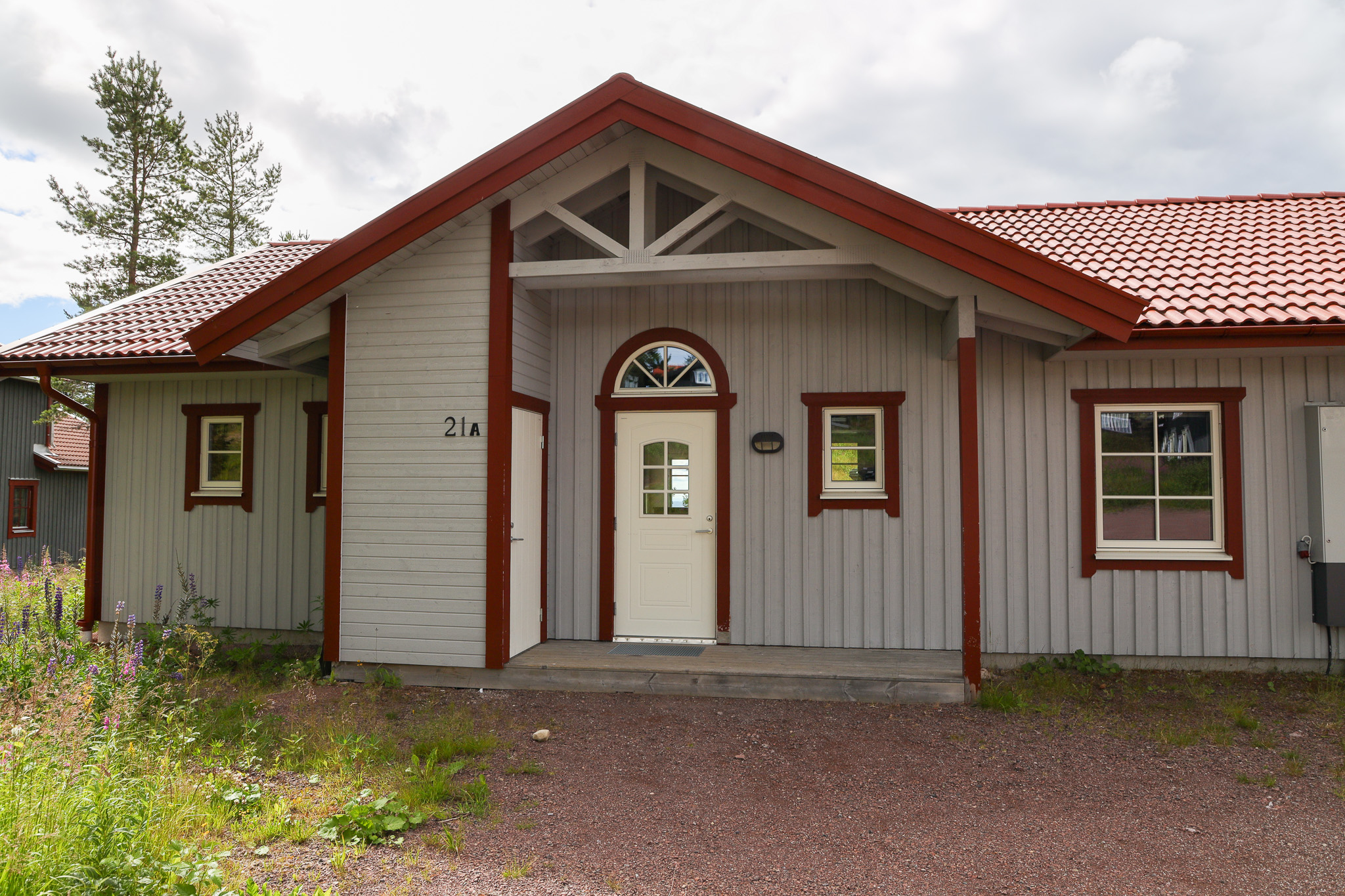 Ljusgrå trähusfasad med rödbruna knutar och rött sadeltak. Entré med vit dörr och fönster ovanför, samt små fönster på båda sidor. Grusgång och gräs i förgrunden, träd i bakgrunden.