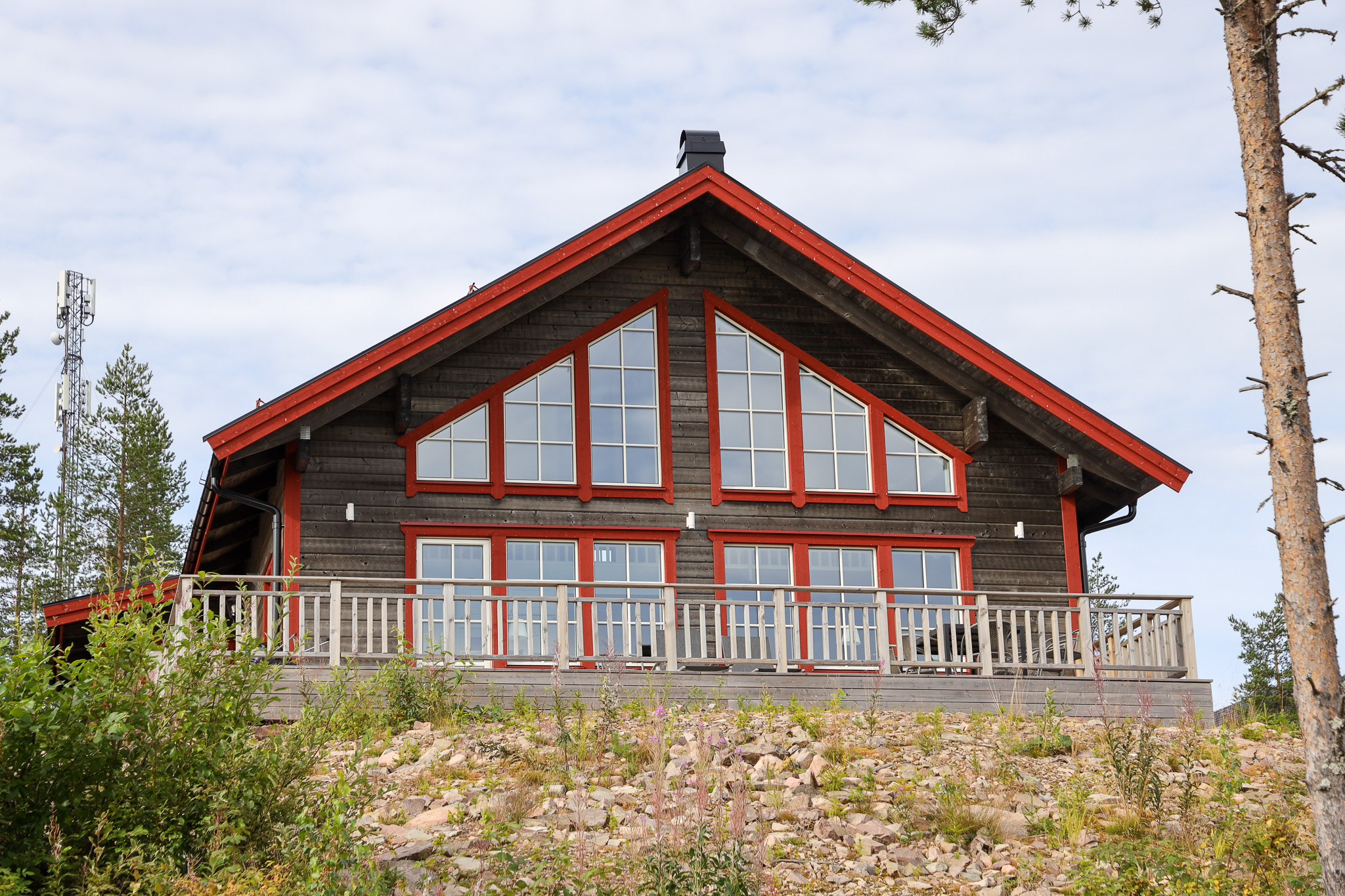 Större timmerstuga med stora fönster och röd takkant, belägen på en liten höjd omgiven av träd.