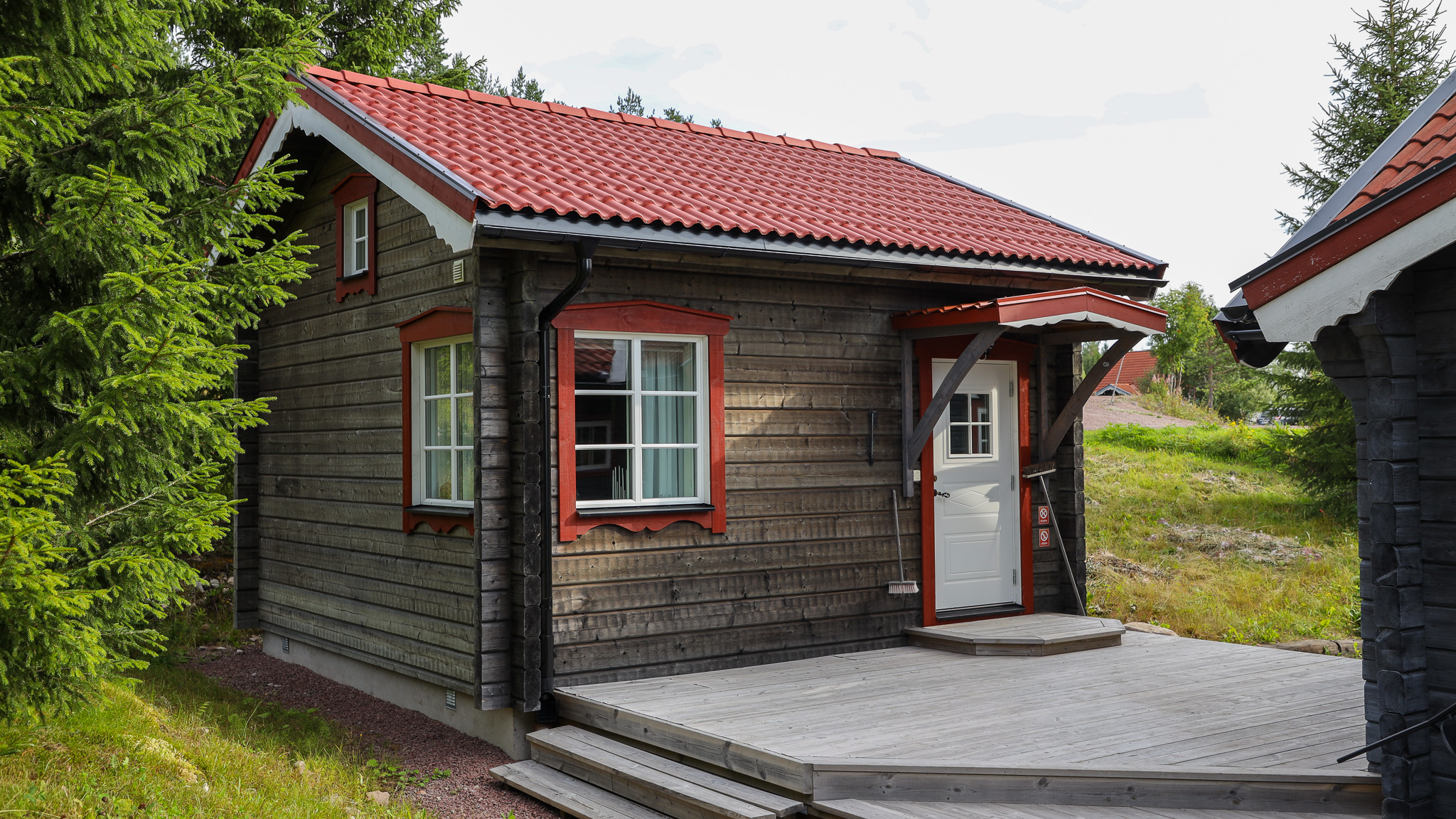 En liten timrad stuga med rött tak och vit dörr, omgiven av gräs och träd i somrig miljö.