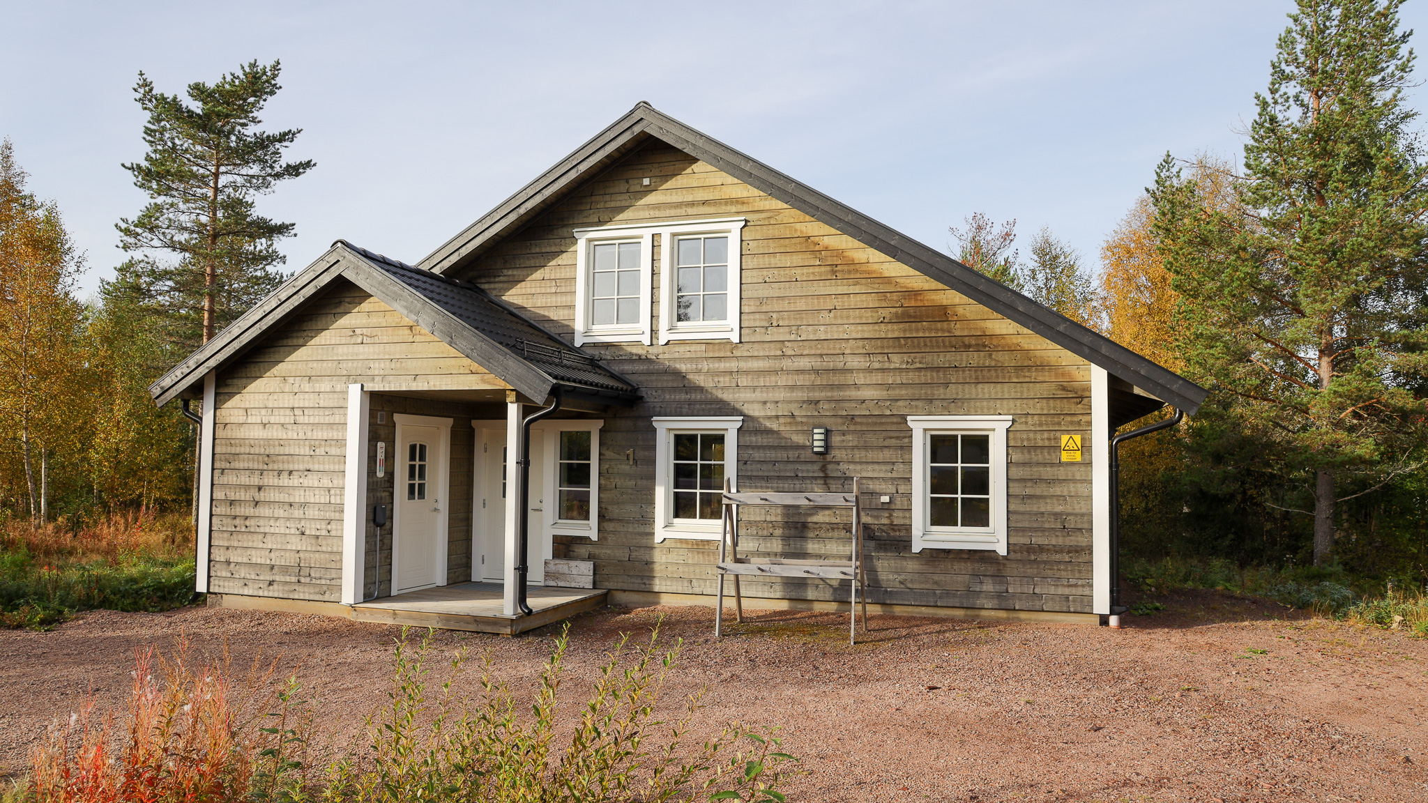Ljusgrå trävilla med vit dörr, vita fönster och sadeltak. Trappa leder upp till en liten balkong på övervåningen. Grusplan framför huset och träd med höstlöv i bakgrunden.