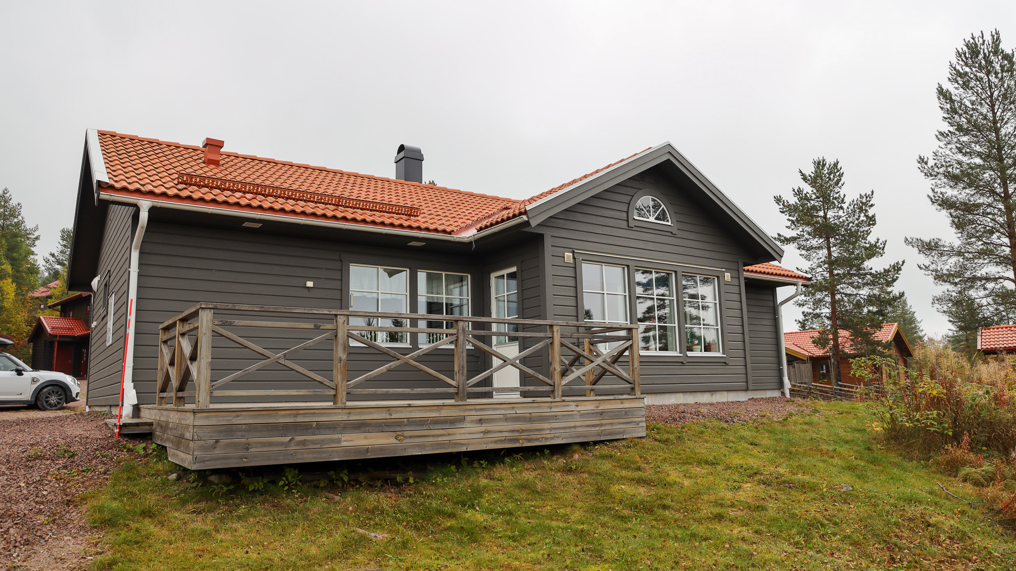 En modern grå trästuga med rött tak och altan, belägen på gräsmark med träd i bakgrunden.