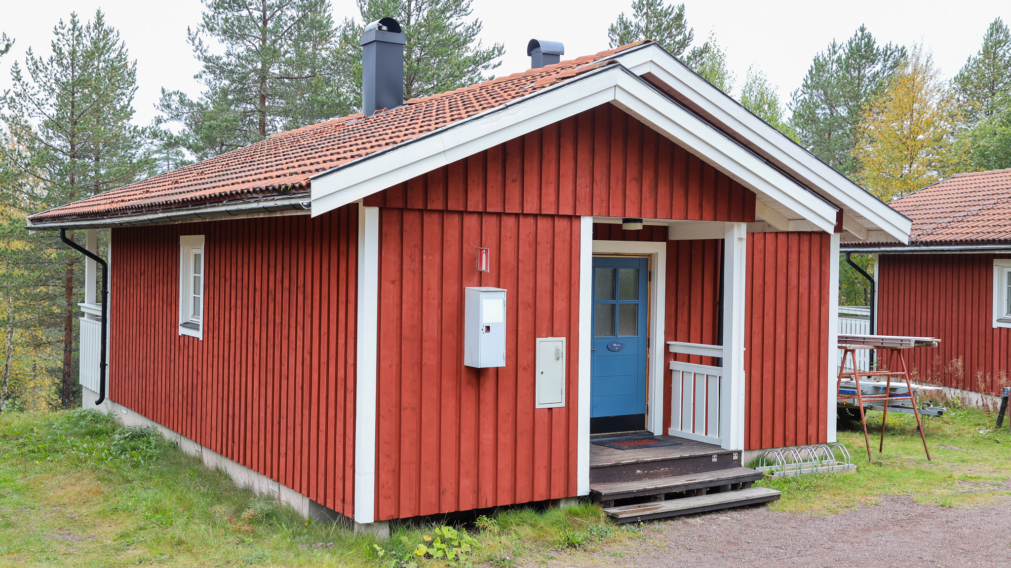 Röd enplansstuga med vita knutar och blå dörr, omgiven av träd och gräsmatta under barmarksperiod.