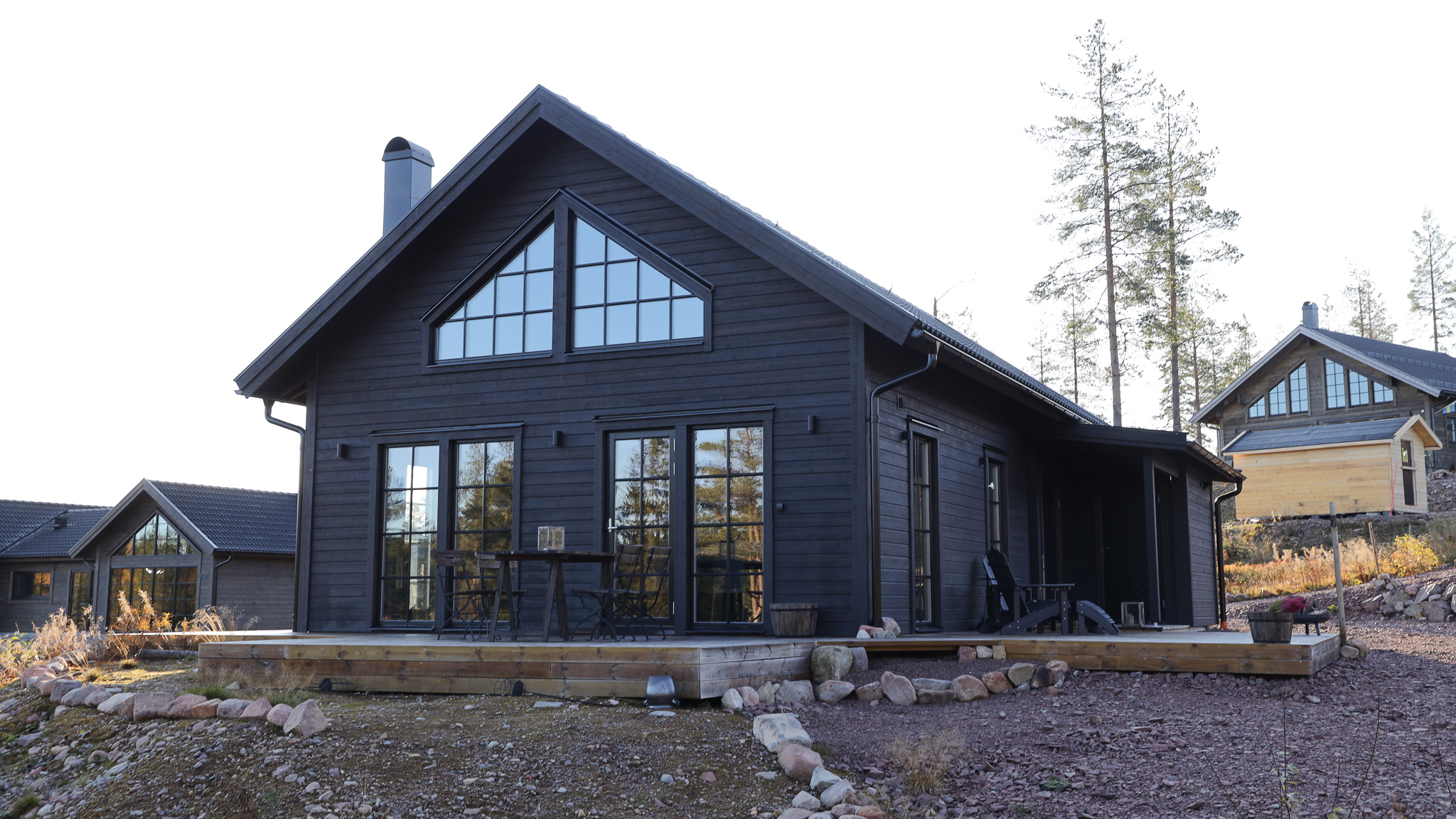 Svartmålad modern fjällstuga med stora fönster och sadeltak, sedd från utsidan i dagsljus. Träterrass framför huset och tallar i bakgrunden.