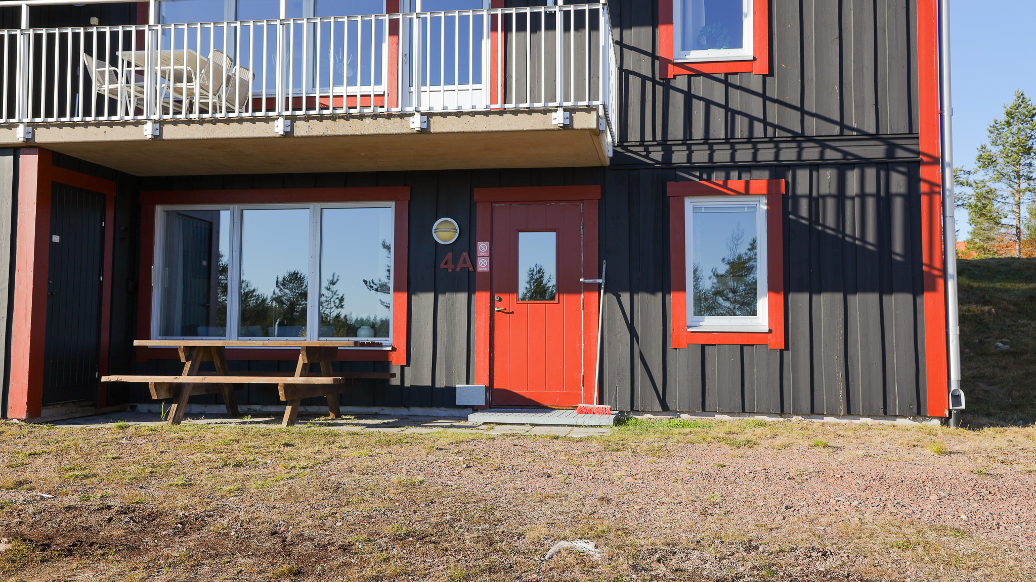 Mörkgrå träbyggnad med röda detaljer runt dörr och fönster. Balkong ovanför entrén och stora glaspartier bredvid dörren. Träbänk framför huset på grusplan.