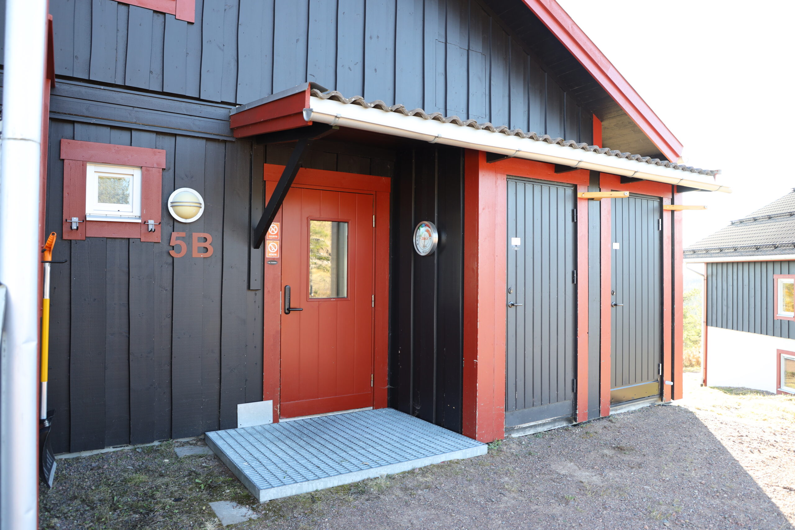 En mörk träbyggnad med röd dörr och detaljer, liten takskyddad entré och grus framför huset.