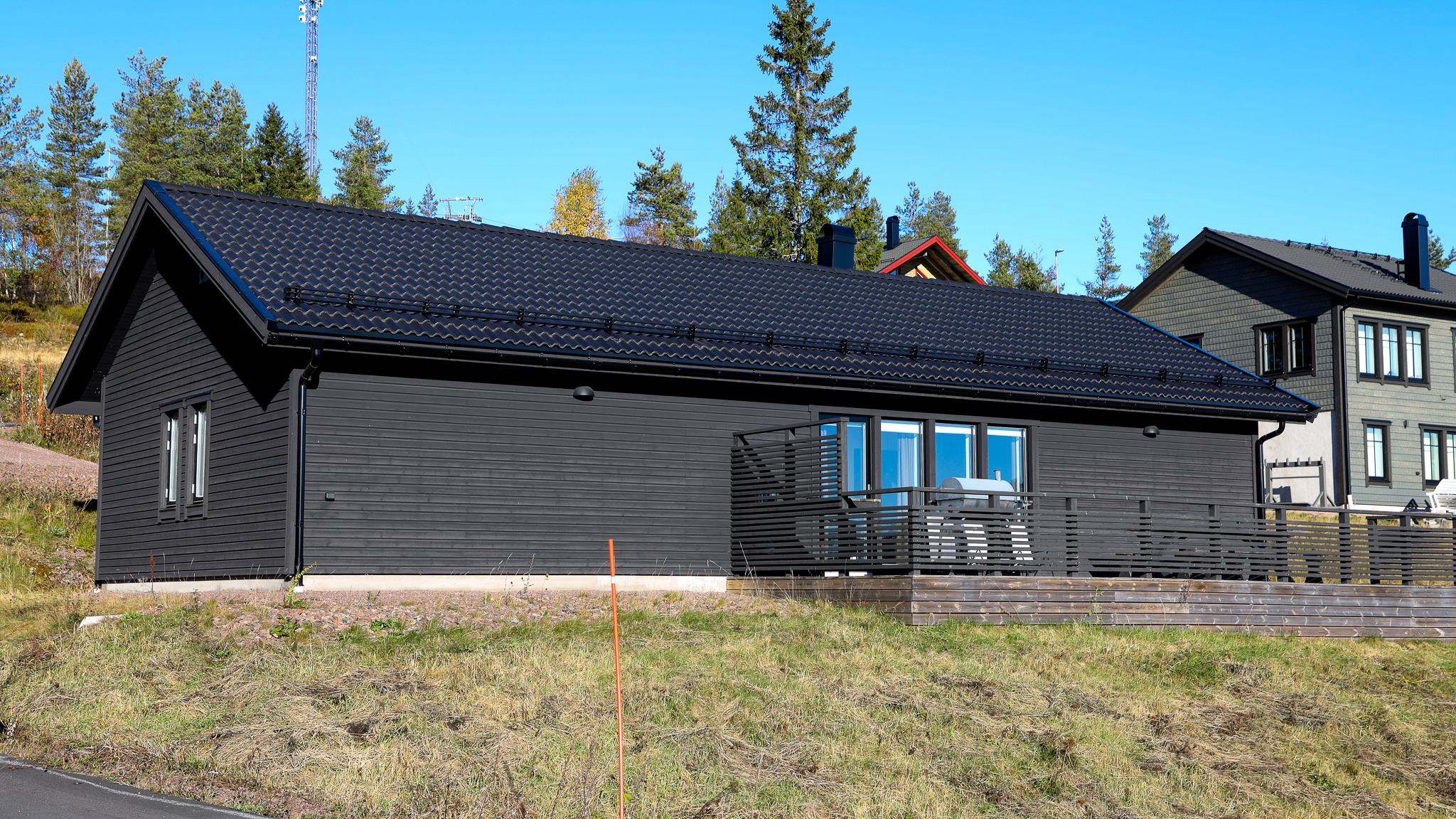 En modern mörk träbyggnad med blå fönsterkarmar och altan, belägen på gräsmark med träd i bakgrunden.