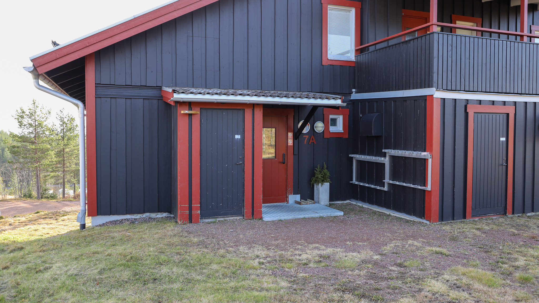 Mörkgrå träbyggnad med röda detaljer runt dörrar och fönster. Ingång med liten trappa och blomkruka vid dörren. Gräs och grus framför huset, träd i bakgrunden.