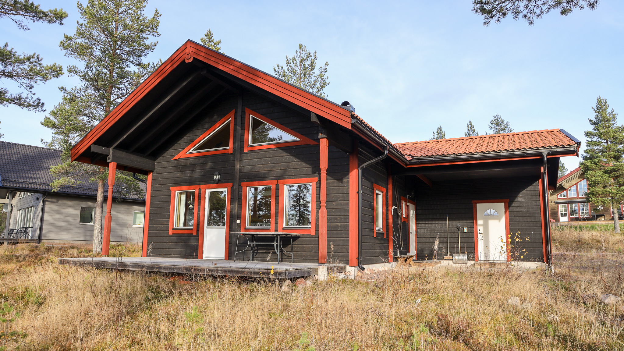 En mörk trästuga med röda detaljer, stora fönster och altan, omgiven av gräs och tallar i soligt väder.