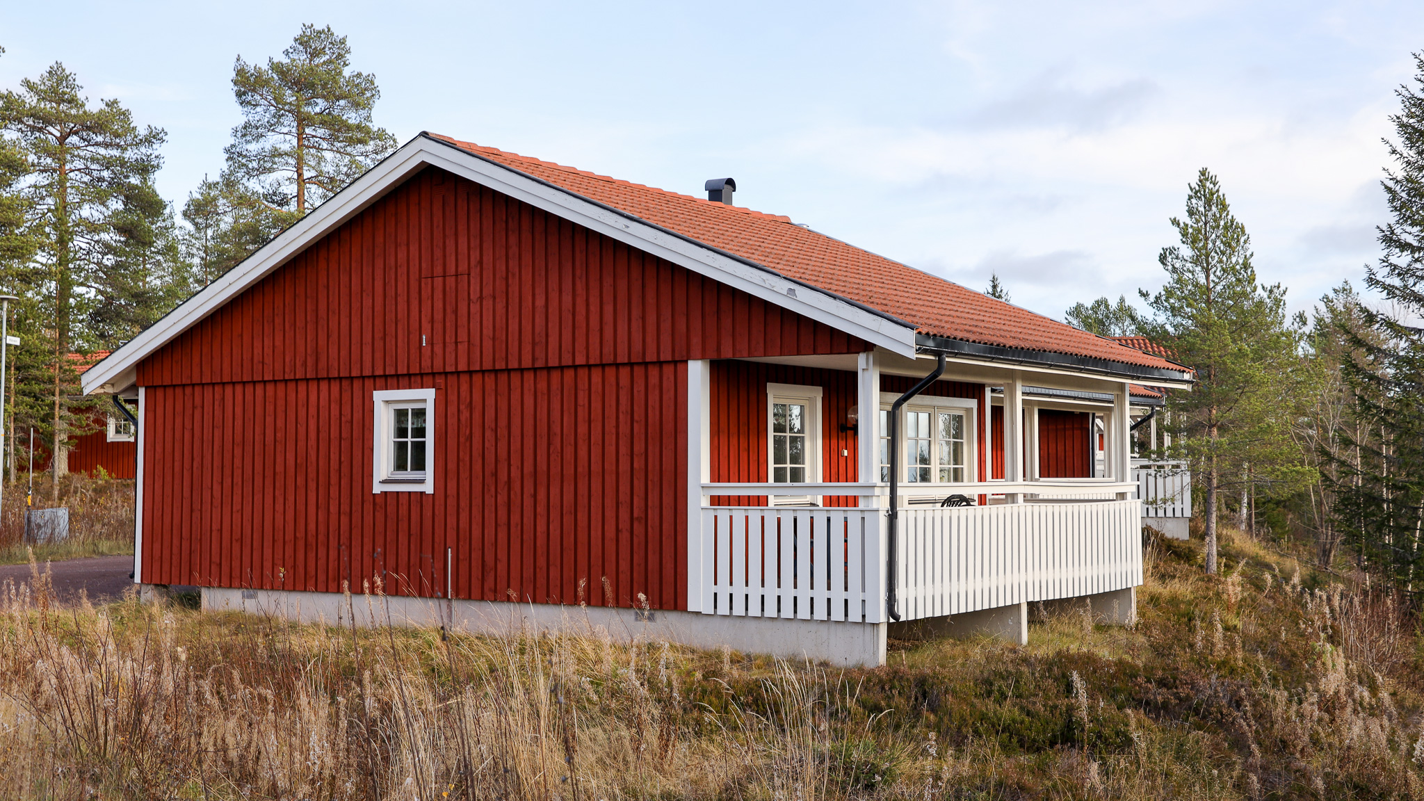 En röd trästuga med vitt räcke och rött tak, omgiven av gräs och tallar i höstmiljö.