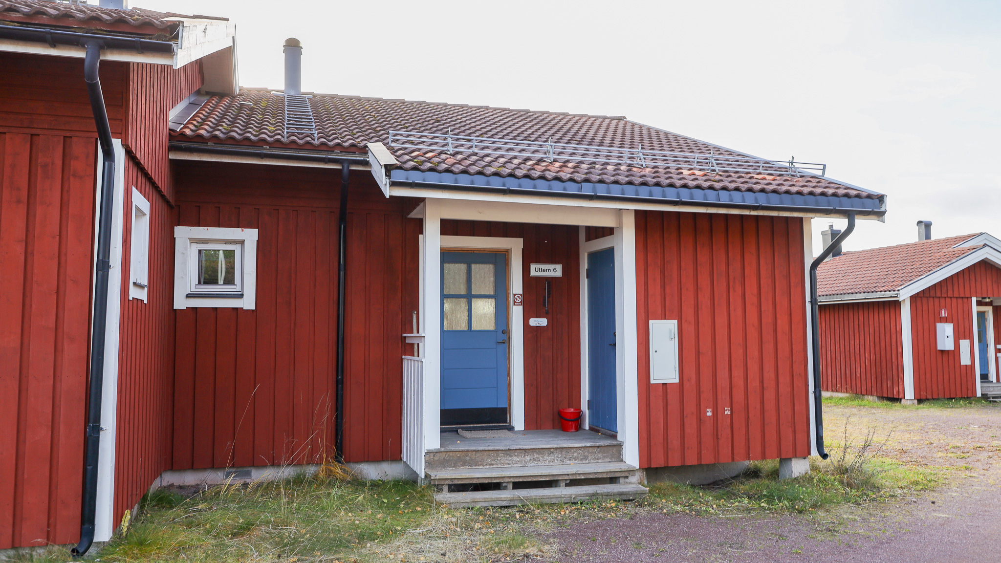 Röd trästuga med vitt takutsprång och blå entrédörr. Liten trappa leder upp till dörren och grusgång syns i förgrunden. Fler röda hus i bakgrunden.