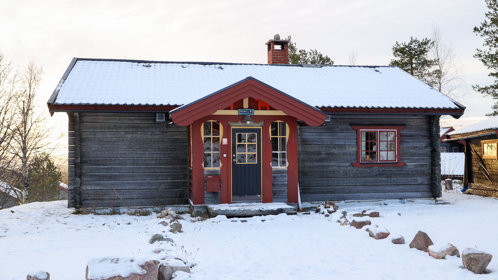 En timrad stuga med röd dörr och snötäckt tak, omgiven av lätt snötäckt mark och träd i bakgrunden.