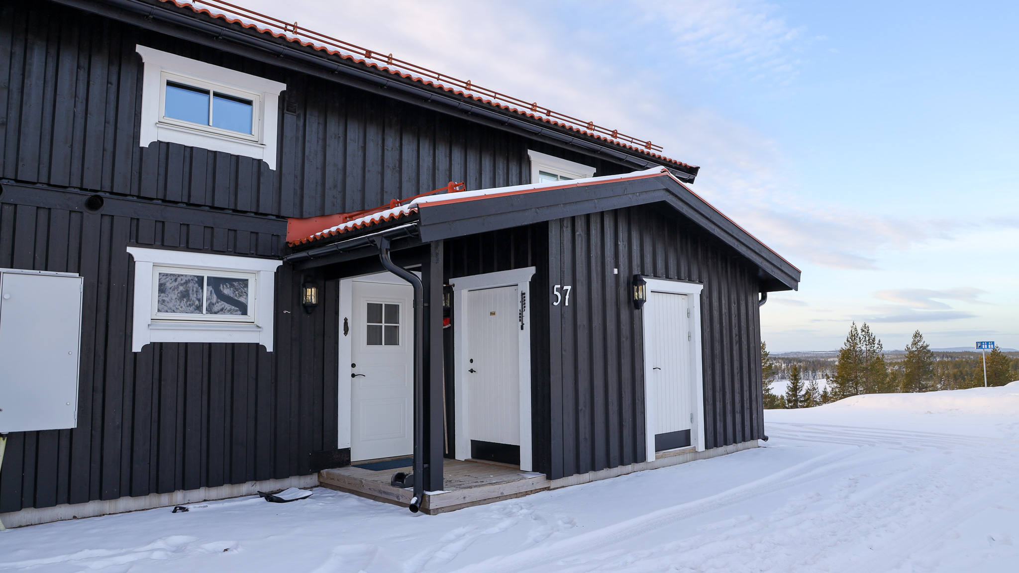 Mörkgrå tvåplansstuga med vit entré och snötäckt mark, med utsikt mot skog och fjäll i bakgrunden.