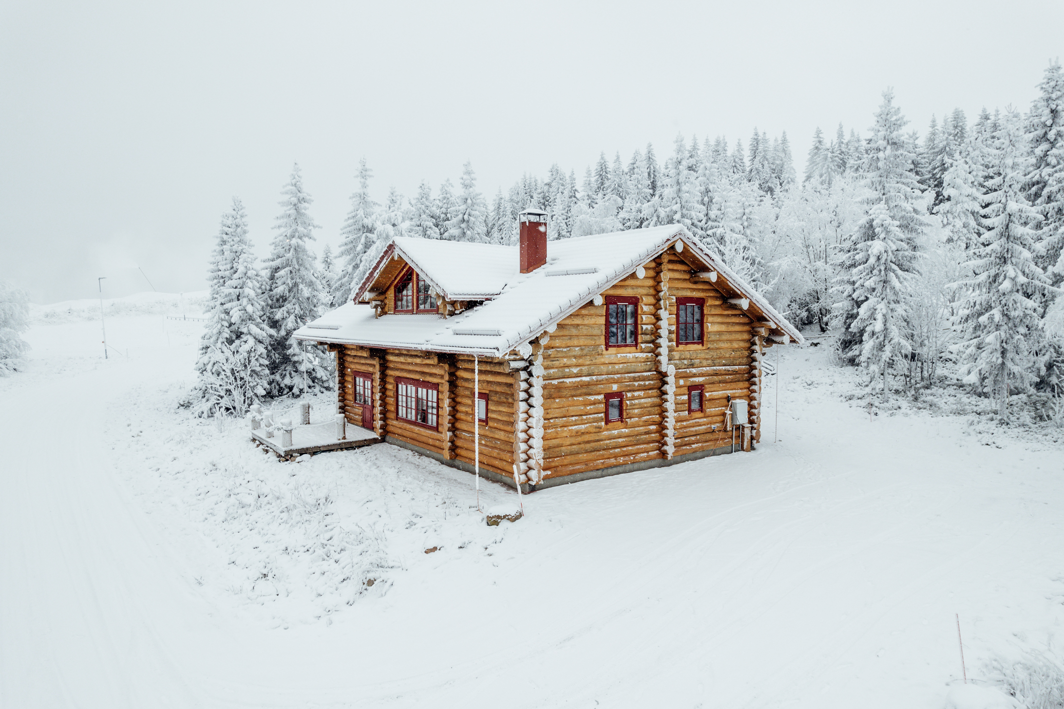 En timrad stuga med snötäckt tak och skorsten, omgiven av snö och barrträd i vinterlandskap.