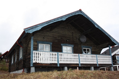 En timrad stuga med blå detaljer, vit veranda och sadeltak, belägen på en gräsbevuxen sluttning.
