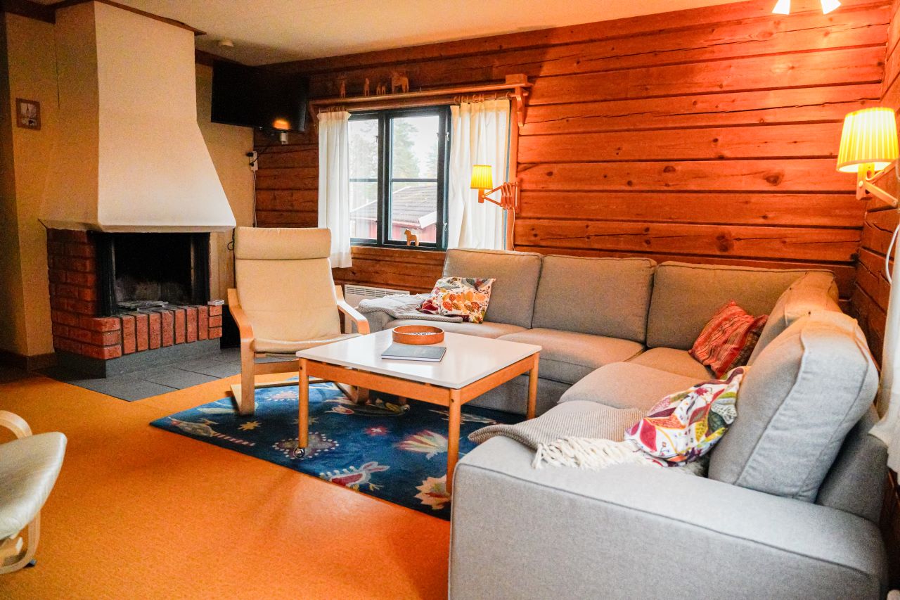 Ett vardagsrum med grå hörnsoffa, vit fåtölj, träbord och öppen spis, med timmerväggar och mysig belysning.