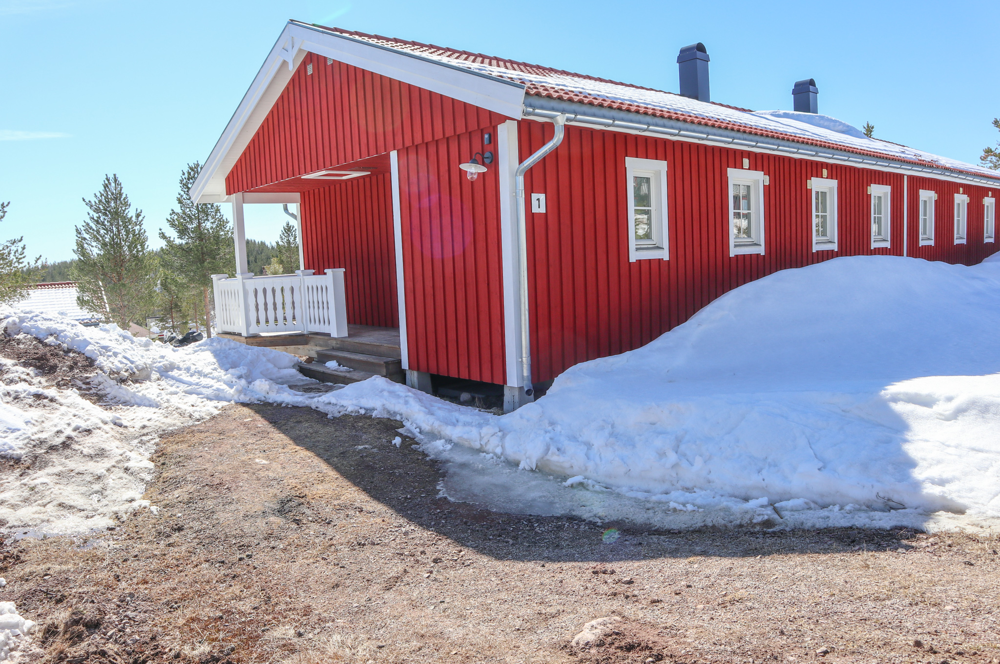 En lång röd trästuga med vit veranda och snö runtomkring, belägen under klarblå himmel.