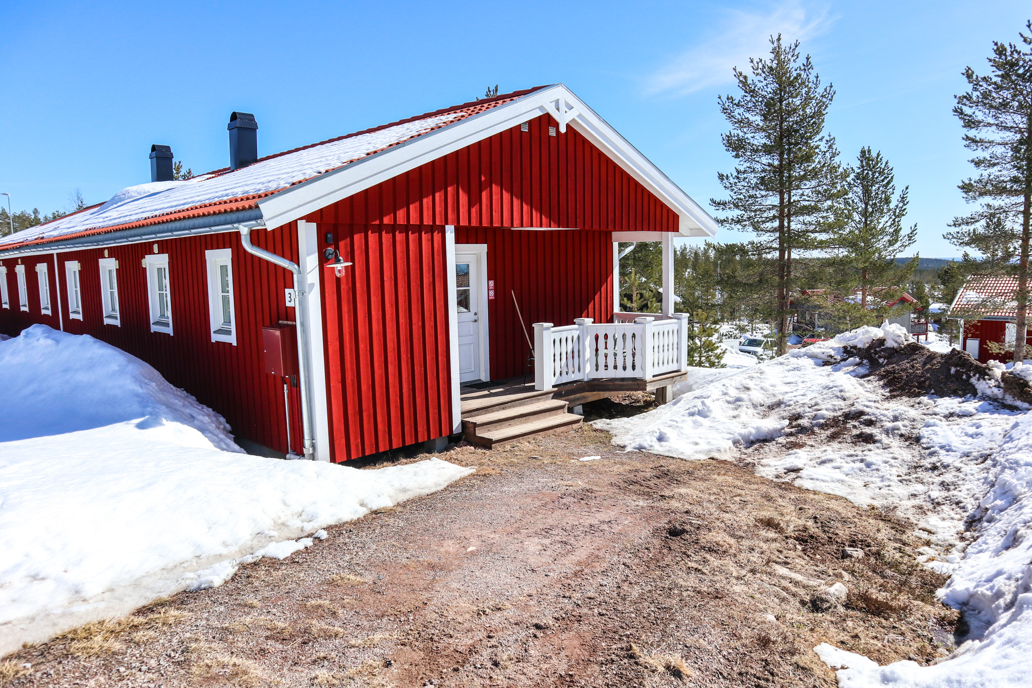 En röd trästuga med vit veranda och snötäckt mark runtom, belägen under klarblå himmel.
