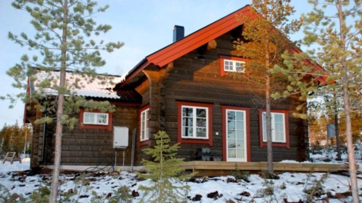 En timrad stuga med röda detaljer och vita fönster, omgiven av snö och tallar i vintermiljö.