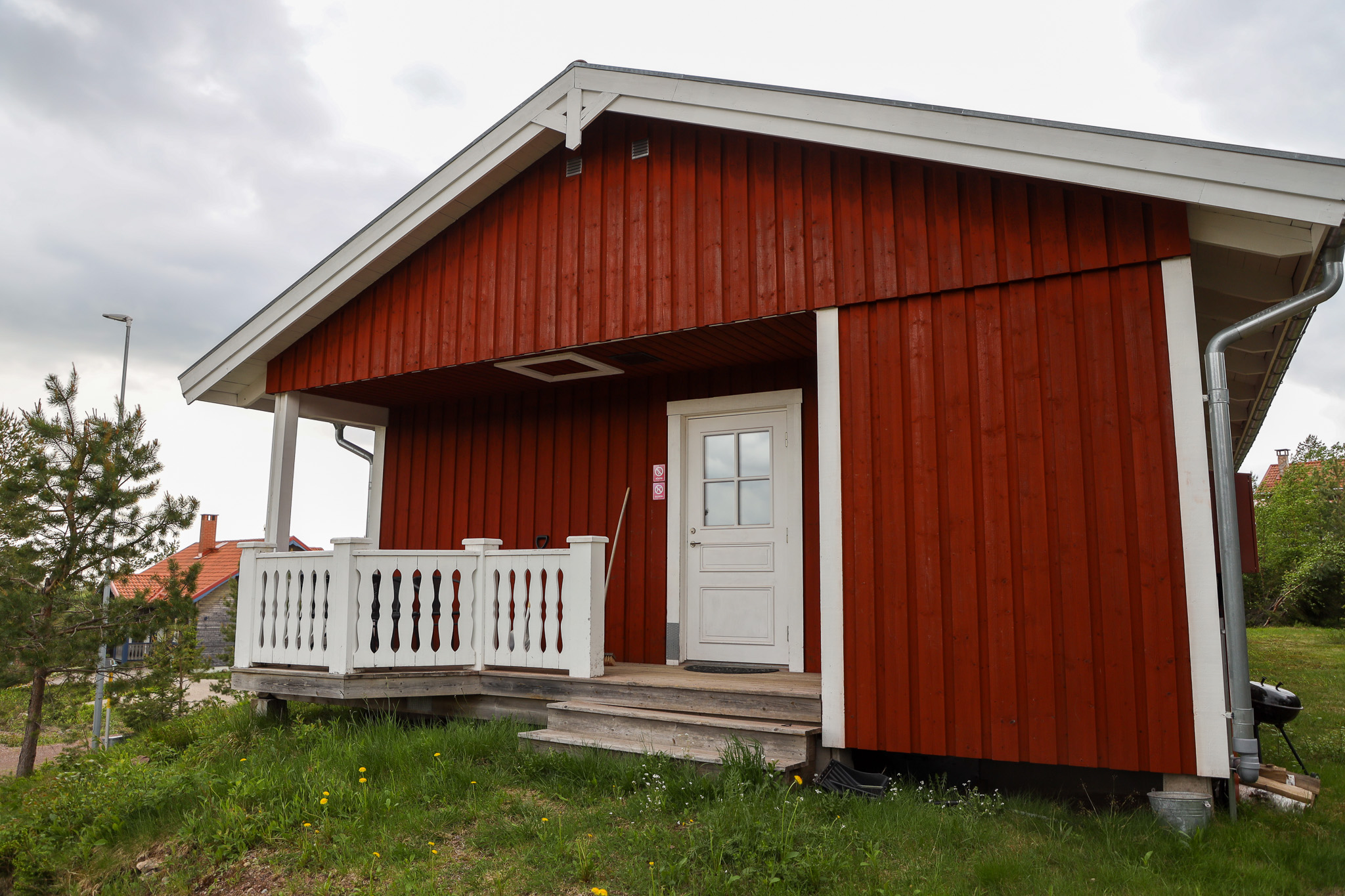 En röd trästuga med vitt räcke och rött tak, omgiven av gräs och tallar i höstmiljö.