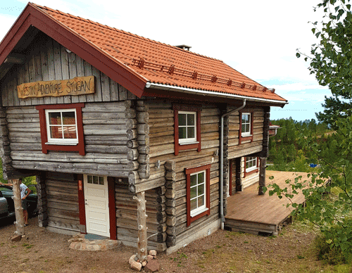Tvåvånings timmerhus med rött tegeltak och röda fönsterkarmar. Vit ytterdörr på nedervåningen och träaltan på sidan av huset. Grus och grönska runt byggnaden.