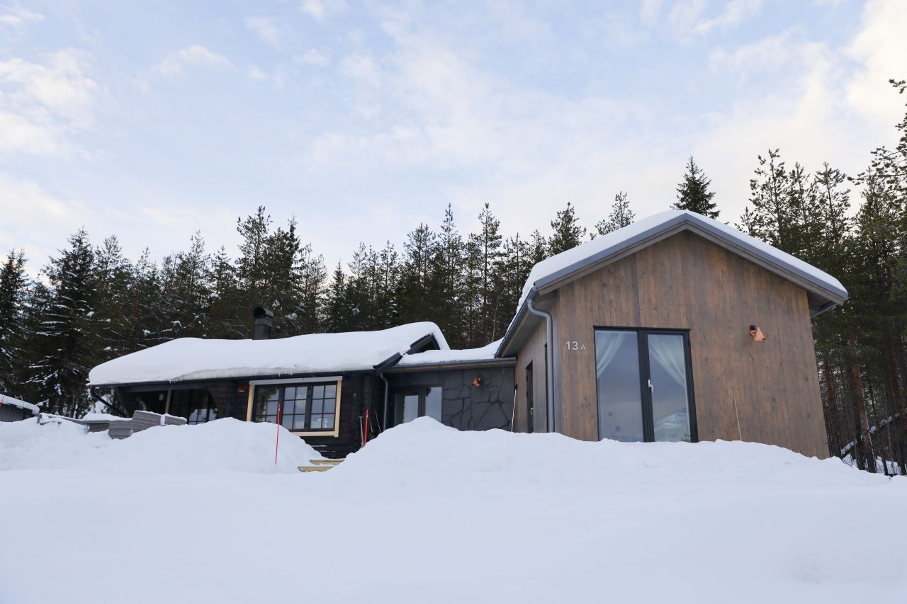 En modern trästuga med stora fönster och snötäckt tak, omgiven av djup snö och tallskog i bakgrunden.