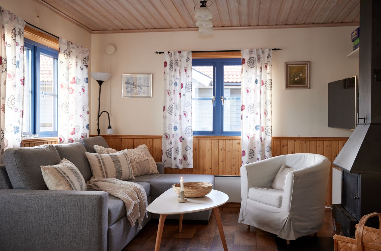 Ett ljust vardagsrum med grå soffa, vit fåtölj, träbord och fönster med blommiga gardiner.