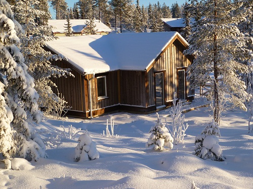 En liten trästuga med snötäckt tak, omgiven av snö och träd i vintermiljö.
