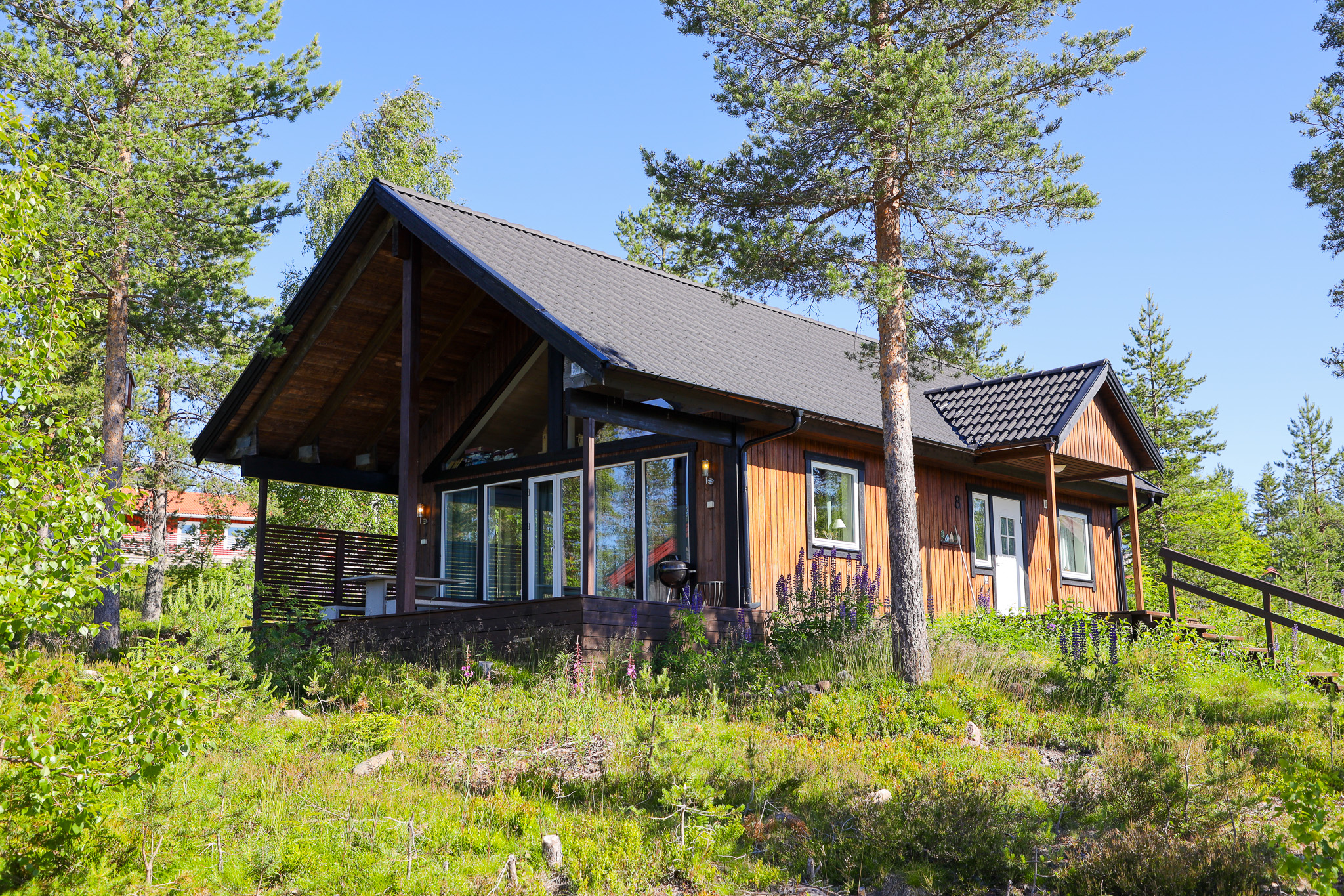 ”Trähus med stora fönster och mörkt sadeltak, omgivet av gräs och tallar i somrig skogsmiljö. Huset är fotat snett framifrån i dagsljus.”