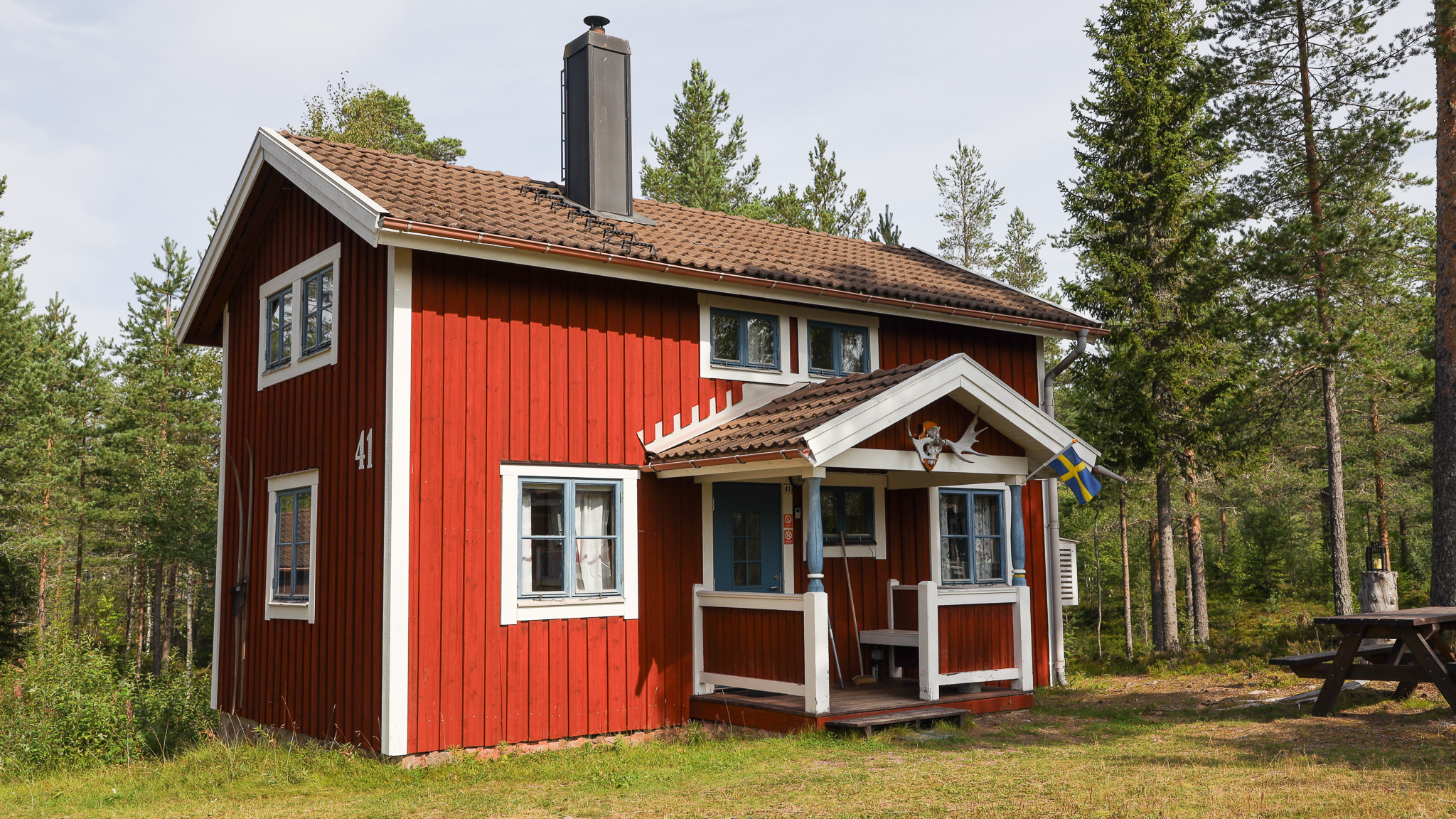 Röd tvåvånings trästuga med vita knutar och liten farstukvist, omgiven av tallar i dagsljus. Ett picknickbord står på marken intill huset.