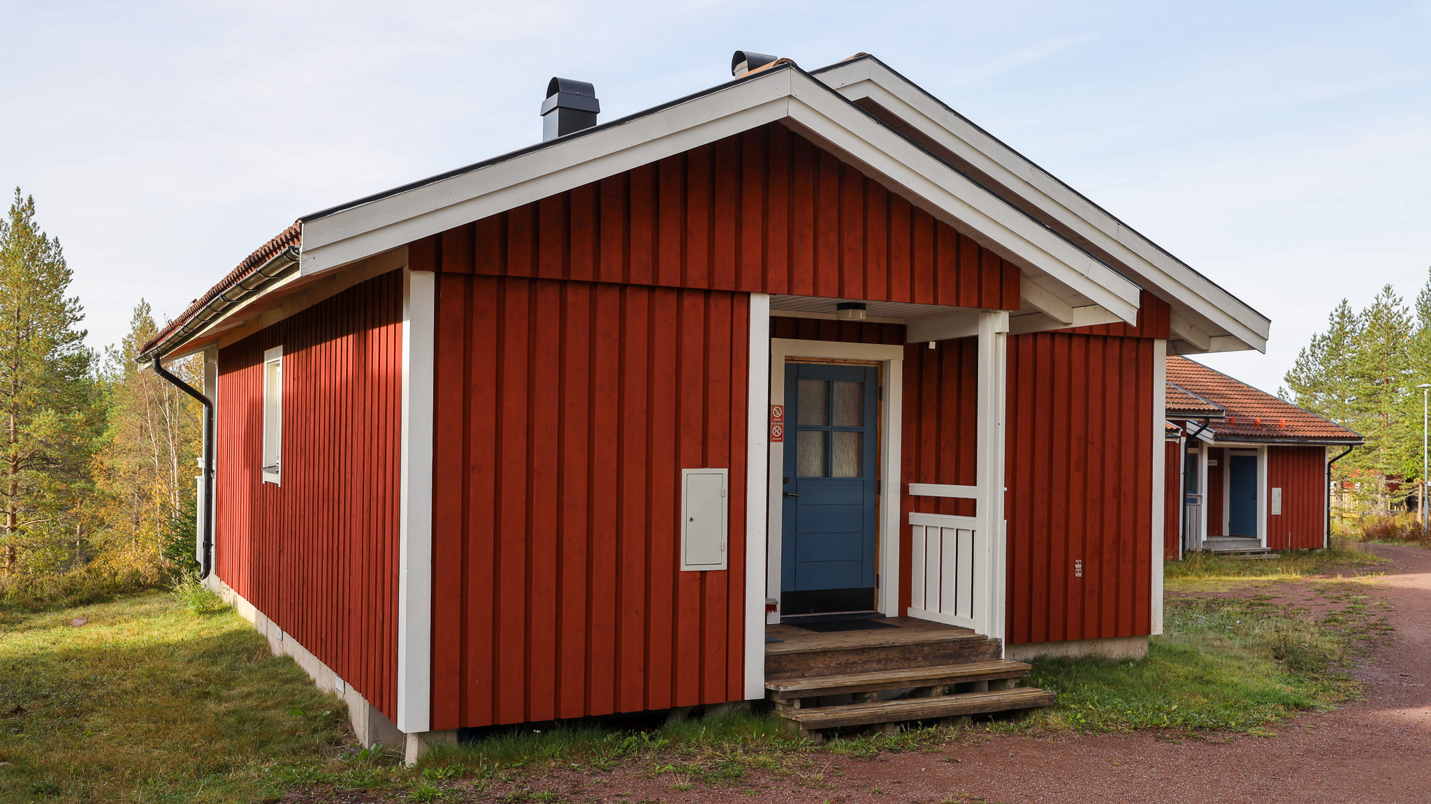 ”Röd trästuga med vita knutar och blå ytterdörr, sedd snett framifrån. Stugan har en liten farstukvist med trappsteg och står i en skogsnära miljö i dagsljus.”