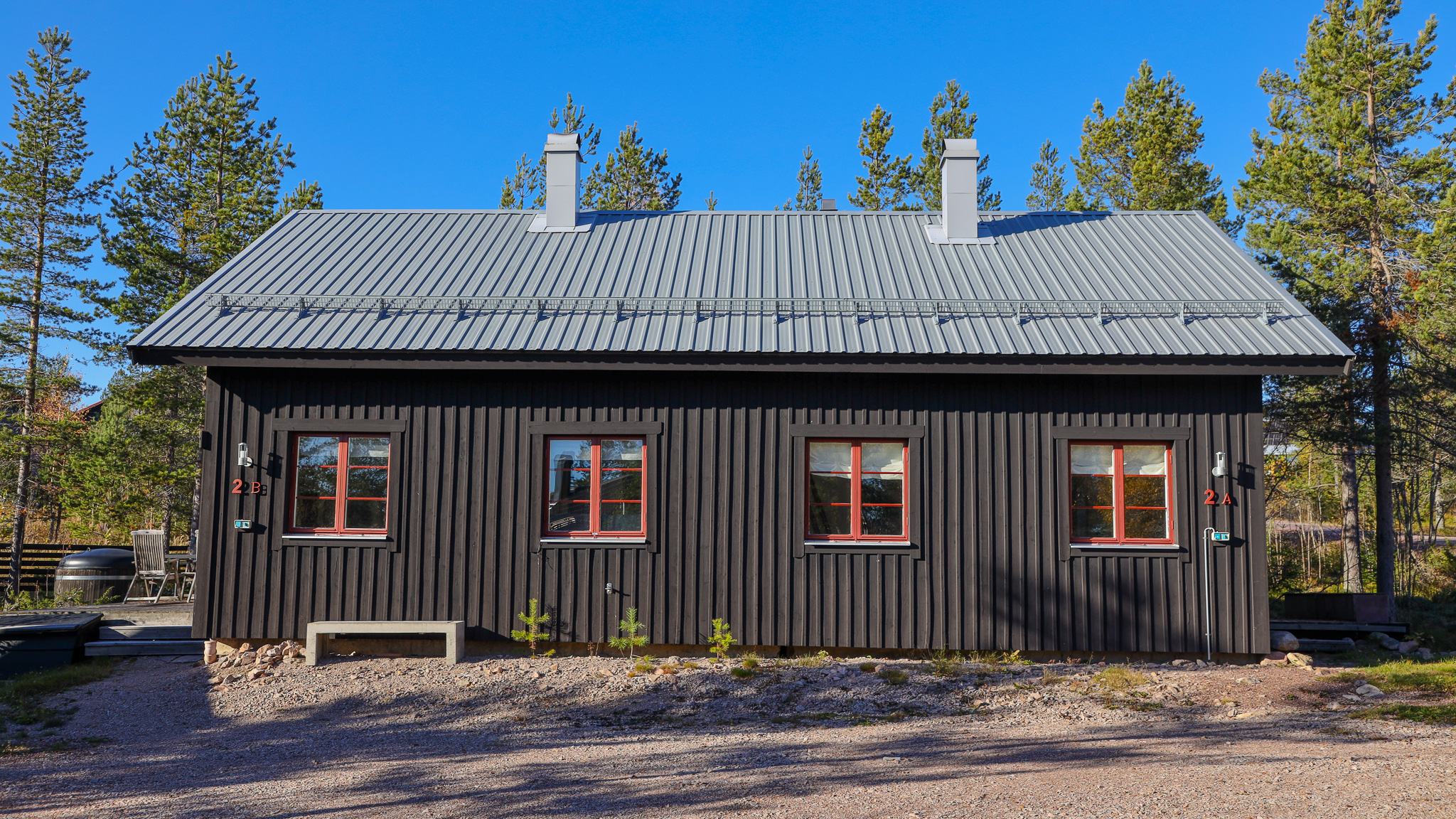 ”Svart timmerbyggnad med grått sadeltak och flera skorstenar, sedd rakt framifrån. Byggnaden står i en tallomgiven miljö i klart dagsljus.”