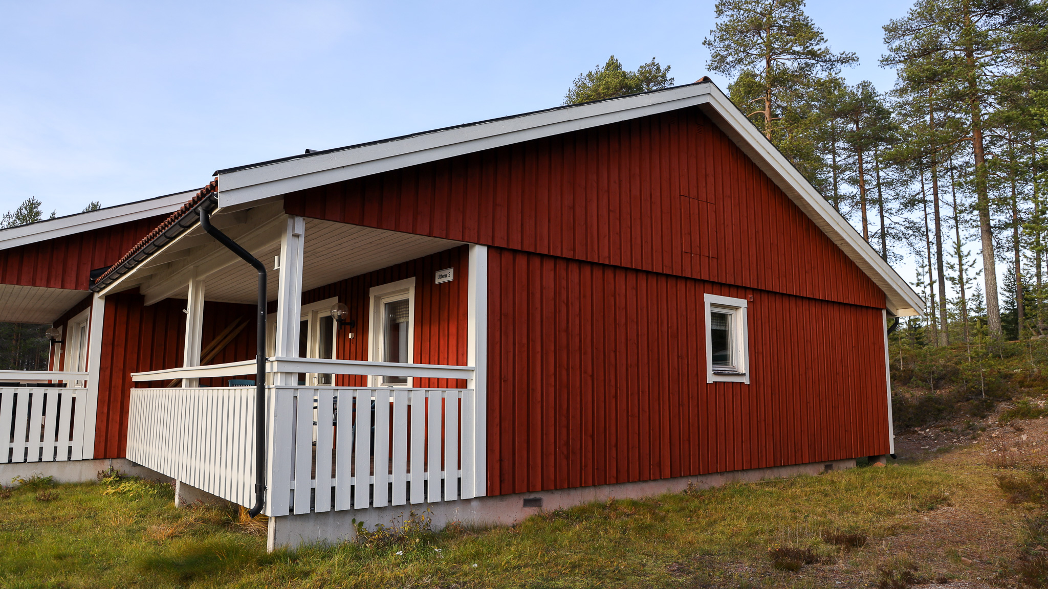 ”Röd enplans trästuga med vita knutar och veranda med räcke, fotad snett framifrån i skogsnära miljö i dagsljus.”