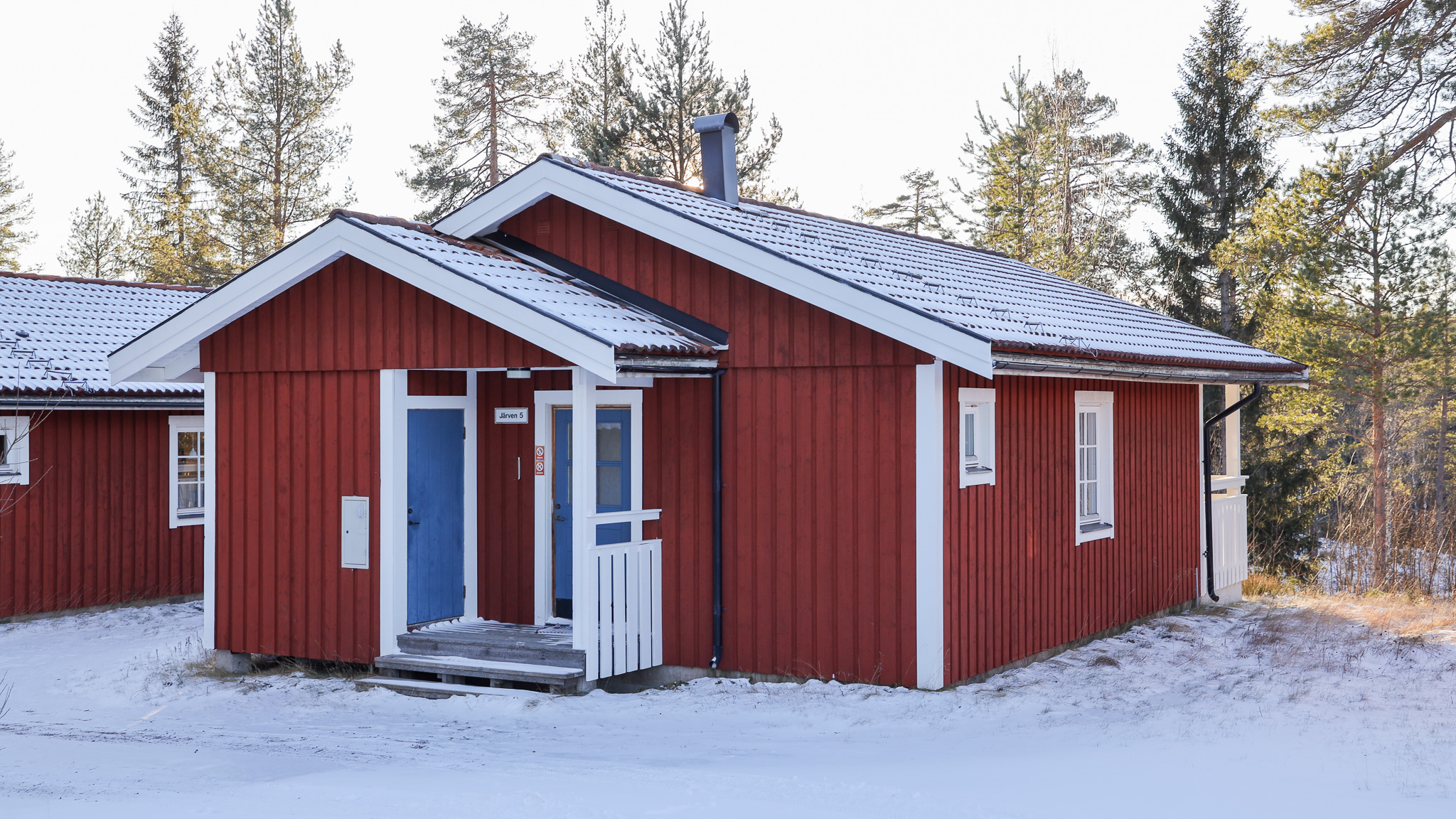 ”Röd enplans trästuga med vita knutar och blå ytterdörr, omgiven av snötäckt mark och tallar. Huset är fotat snett framifrån i vinterljus.”