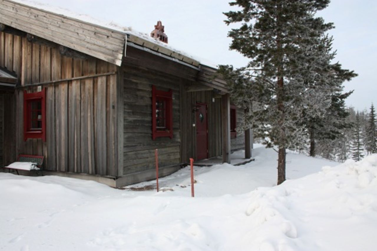”Träbyggnad med grå fasad och röda fönsterkarmar, omgiven av djup snö och tallar. Huset är fotat snett framifrån i vinterljus.”