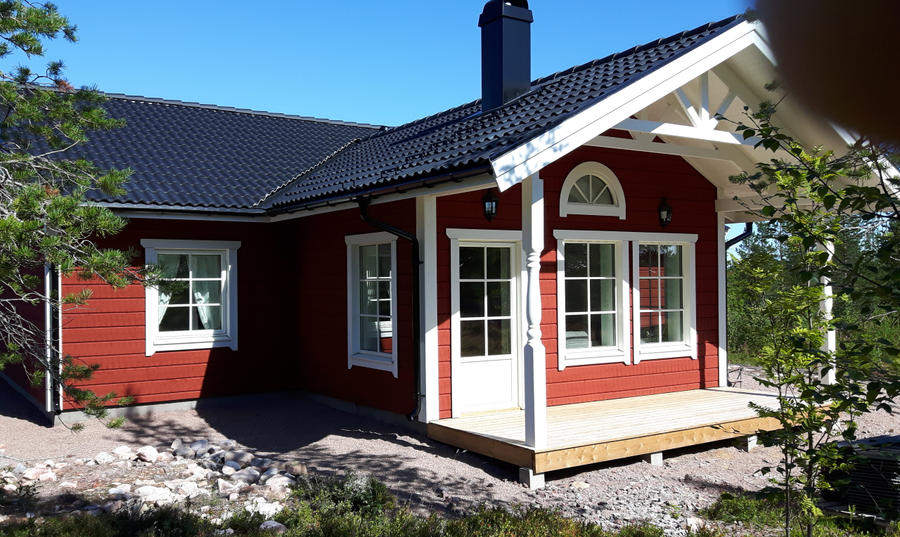”Röd trästuga med vita knutar, stora fönster och mörkt tak, fotad snett framifrån i soligt dagsljus. Huset omges av stenig mark och gles vegetation.