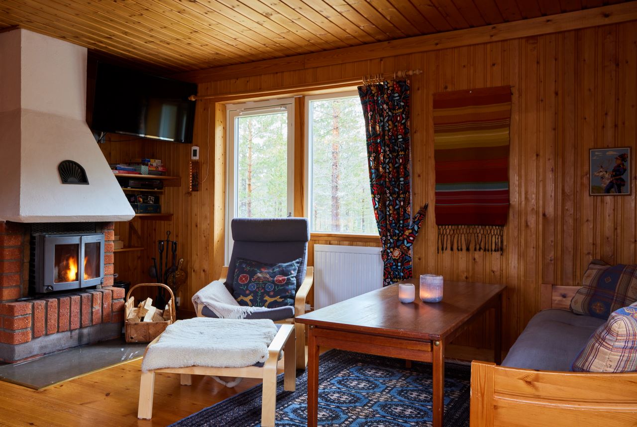 ”Vardagsrum med träpanel, öppen spis, fåtöljer, soffbord och fönster med utsikt mot skog. Rummet är varmt belyst och inrett i rustik stil.”