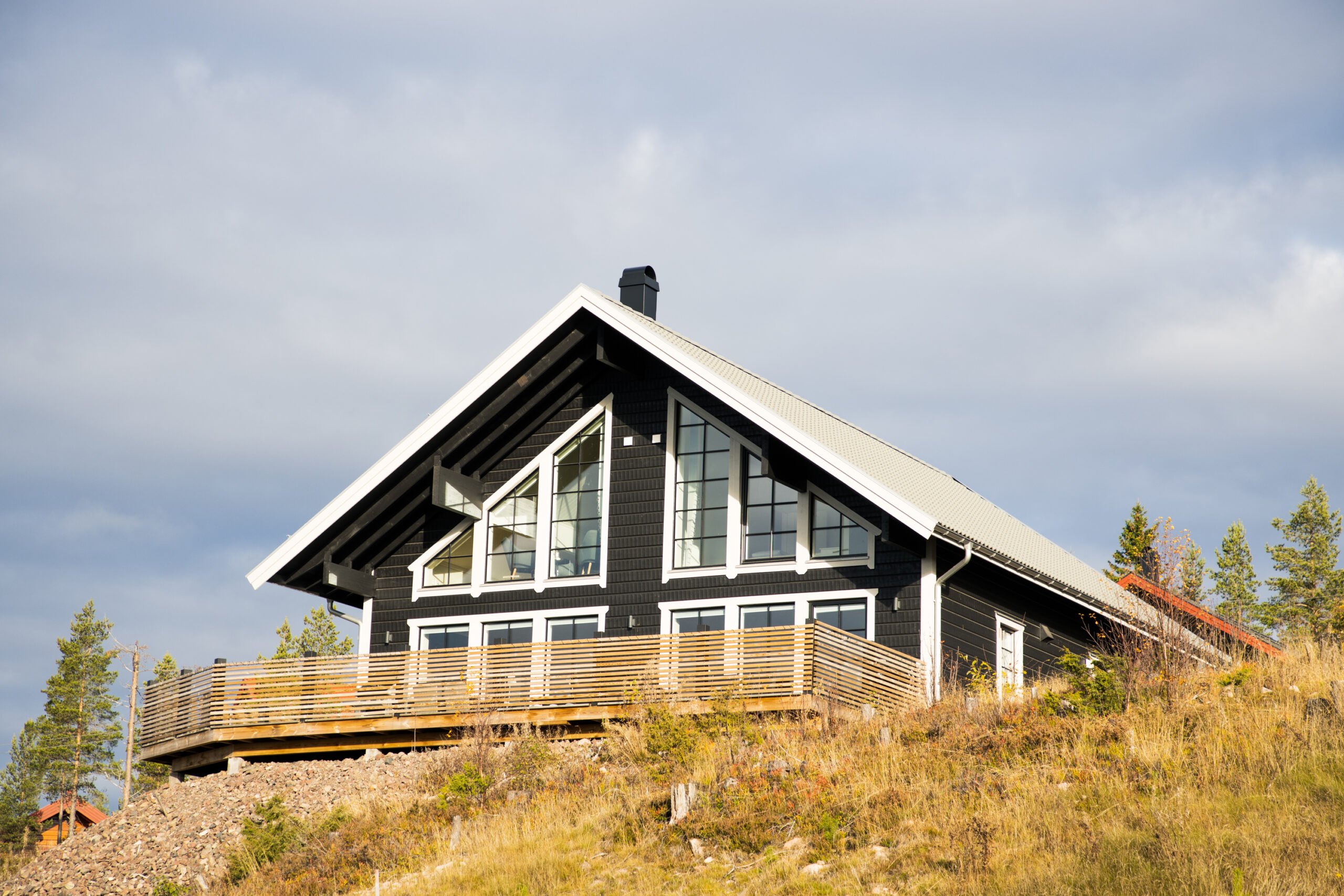 ”Svart trähus med stora fönsterpartier och ljus altan, beläget på en sluttning i öppen natur, fotat framifrån i dagsljus.”