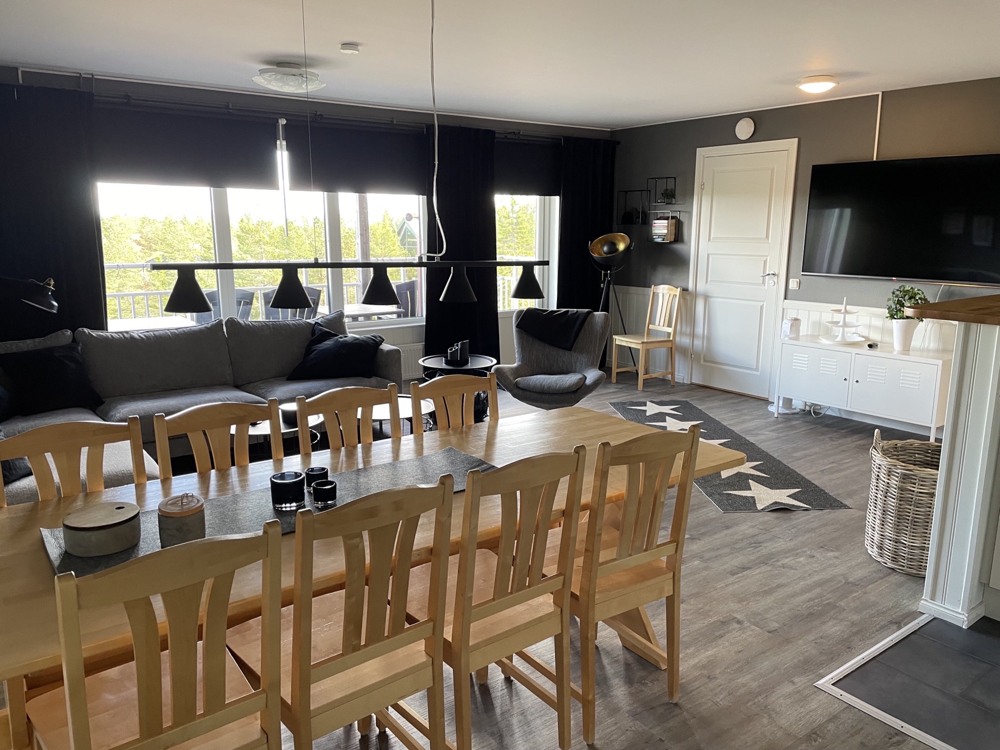 ”Ljust mat- och vardagsrum med stort matbord i trä, soffgrupp vid fönstren, väggmonterad TV och öppet kök. Rummet är inrett i moderna färger och fotat i dagsljus.”
