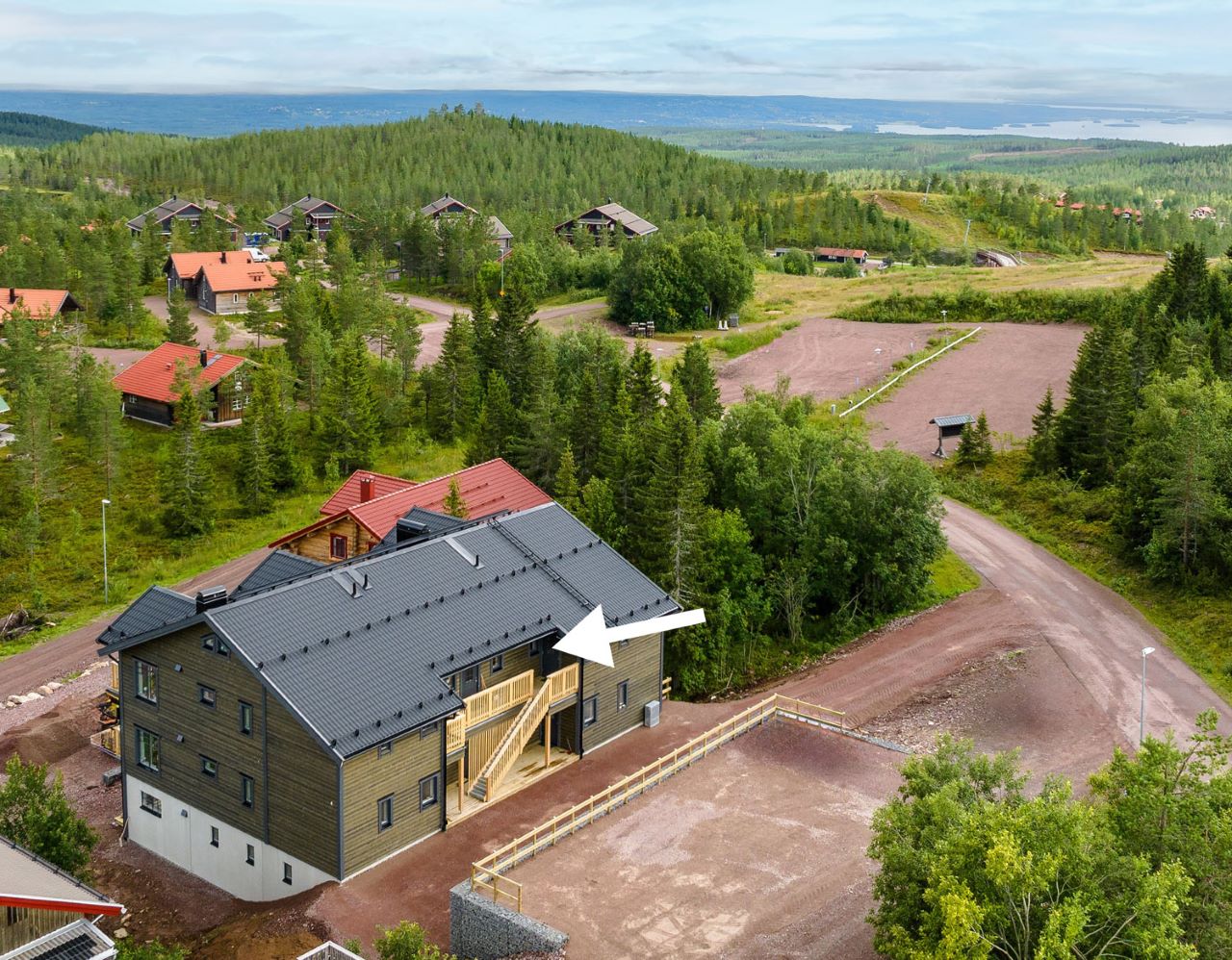 ”Större byggnad med mörkt tak och ljus fasad, fotad från ovan i ett kuperat skogsområde. En vit pil pekar mot byggnadens entré på den högra sidan.”