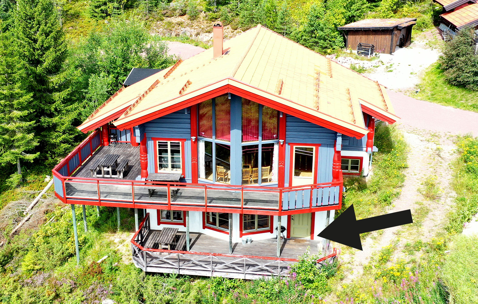 ”Tvåvånings trähus med röd och blå fasad samt rymliga balkonger, fotat från ovan i somrig skogsmiljö. En svart pil pekar mot byggnadens nedre högra del.”