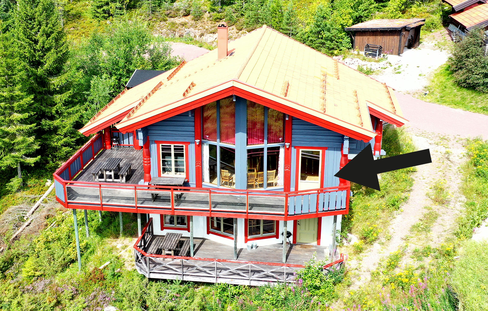 ”Tvåvånings trähus med röd och blå fasad samt stort balkongräcke, fotat från ovan i somrig skogsmiljö. En svart pil pekar mot byggnadens högra del.”