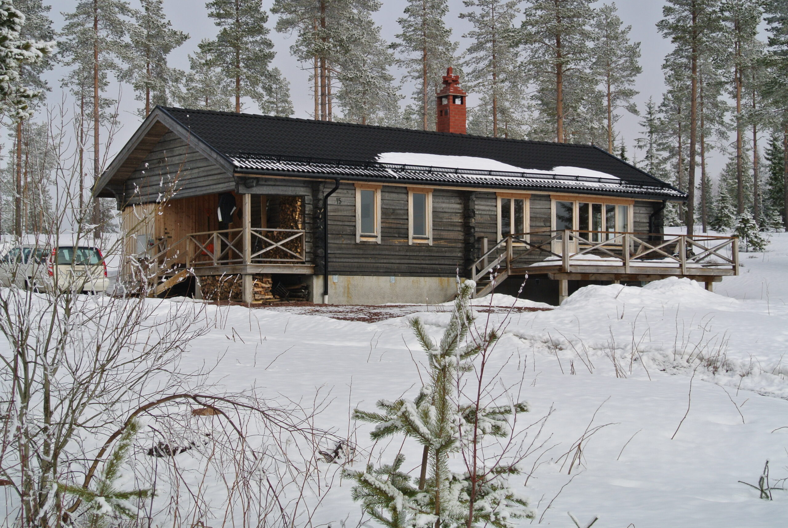 Mörk trähytta med snötäckt tak och veranda, omgiven av tallar och vinterlandskap i Orsa Grönklitt.