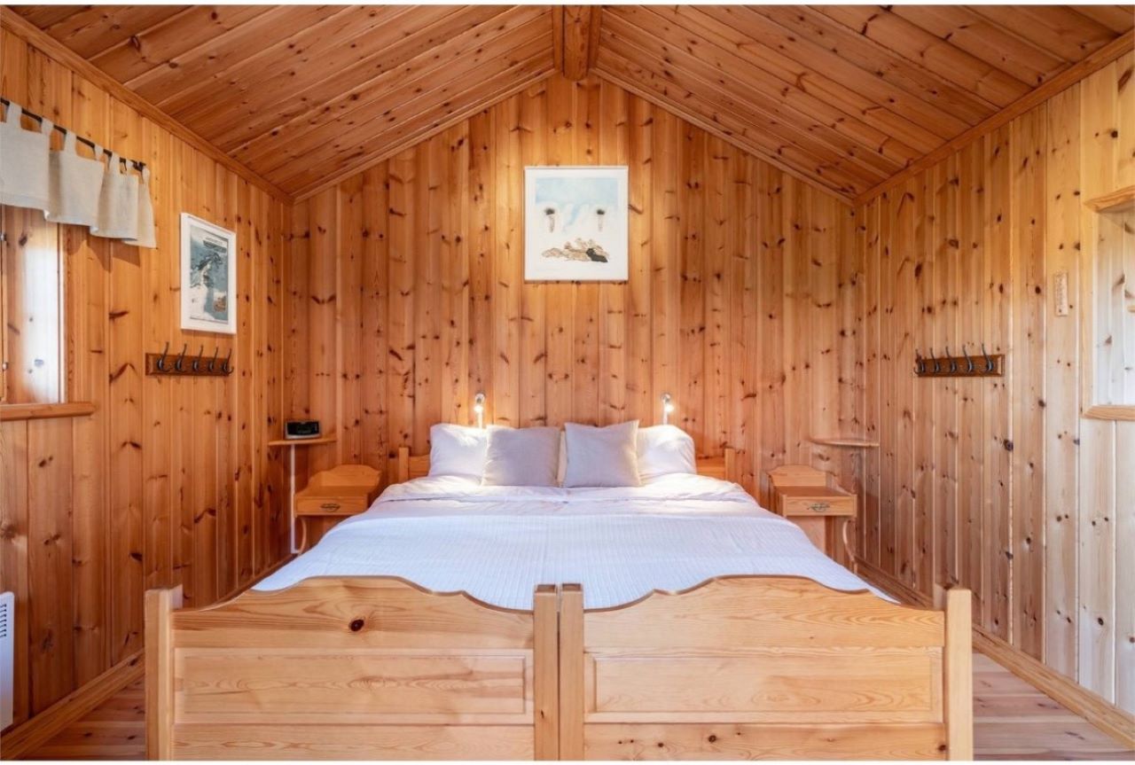 Dubbelsäng i ett ljust sovrum med träpanel, snedtak och fönster i en stuga. Dubbelsäng i ett ljust sovrum med träpanel, snedtak och fönster i en stuga.