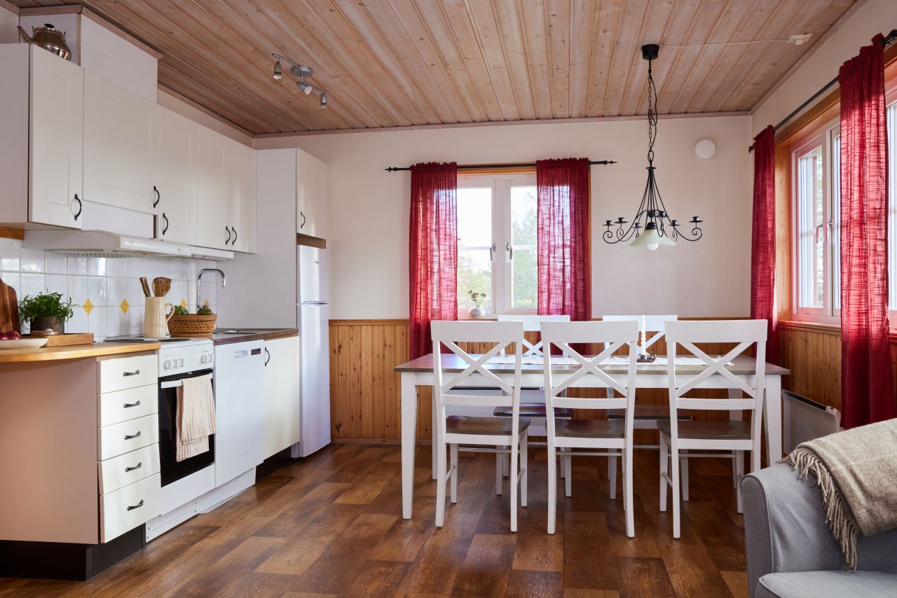 Kök med matplats, matbord och stolar, vita köksskåp och fönster med gardiner.