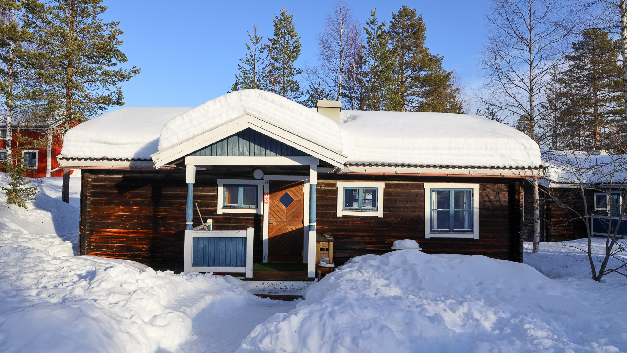 Enplansstuga i trä med snötäckt tak och entré på framsidan, omgiven av snö och träd. Enplansstuga i trä med snötäckt tak och entré på framsidan, omgiven av snö och träd.